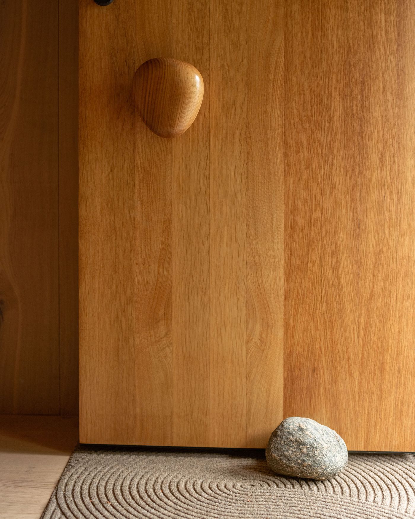 A wooden door with a smooth round handle stands beside a textured rug and a gray stone resting on the floor. A wooden door with a smooth round handle stands beside a textured rug and a gray stone resting on the floor.
