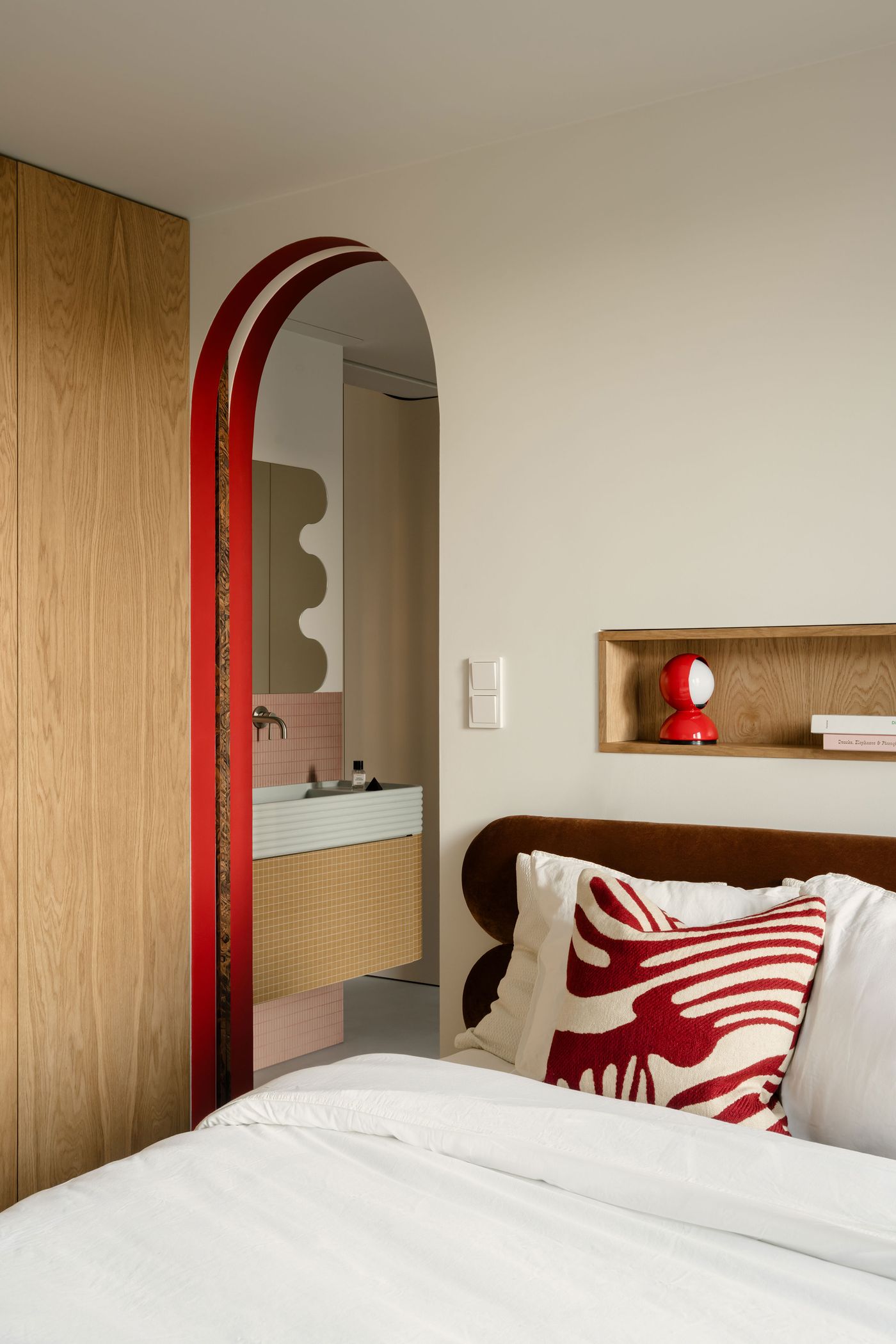 An arched passage with red trim opens into a boldly styled bathroom, seen from the bedroom. The layering of sculptural forms, warm woods, and playful red accents hints at a seamless transition between private and public zones. An arched passage with red trim opens into a boldly styled bathroom, seen from the bedroom. The layering of sculptural forms, warm woods, and playful red accents hints at a seamless transition between private and public zones.