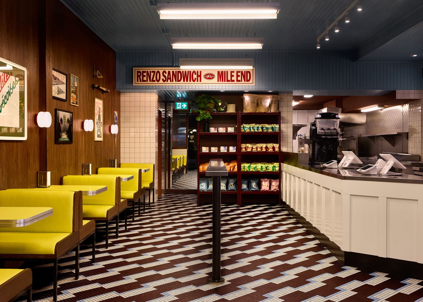 The interior of Renzo Sandwich Shop features a long view of bright yellow booths and tables with chrome trim, set against warm wood-paneled walls. The distinctive retro aesthetic is enhanced by a patterned tiled floor and a prominent "RENZO SANDWICH MILE END" sign. Shelving units display products, and a service counter runs along the right side.