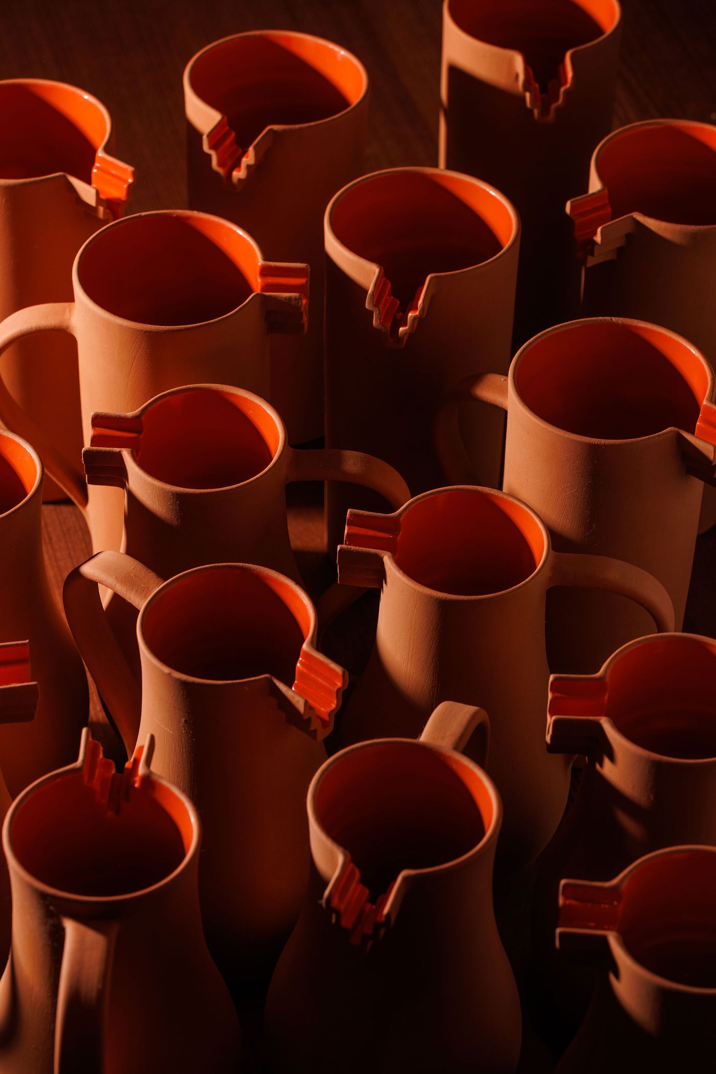 A close-up view of numerous artisanal terracotta pitchers, each featuring a bright orange interior and a uniquely notched spout, arranged in a textured pattern. The warm, earthy tones of the pottery are beautifully illuminated, highlighting their handcrafted quality and the subtle play of light and shadow, evoking a rustic yet refined aesthetic.