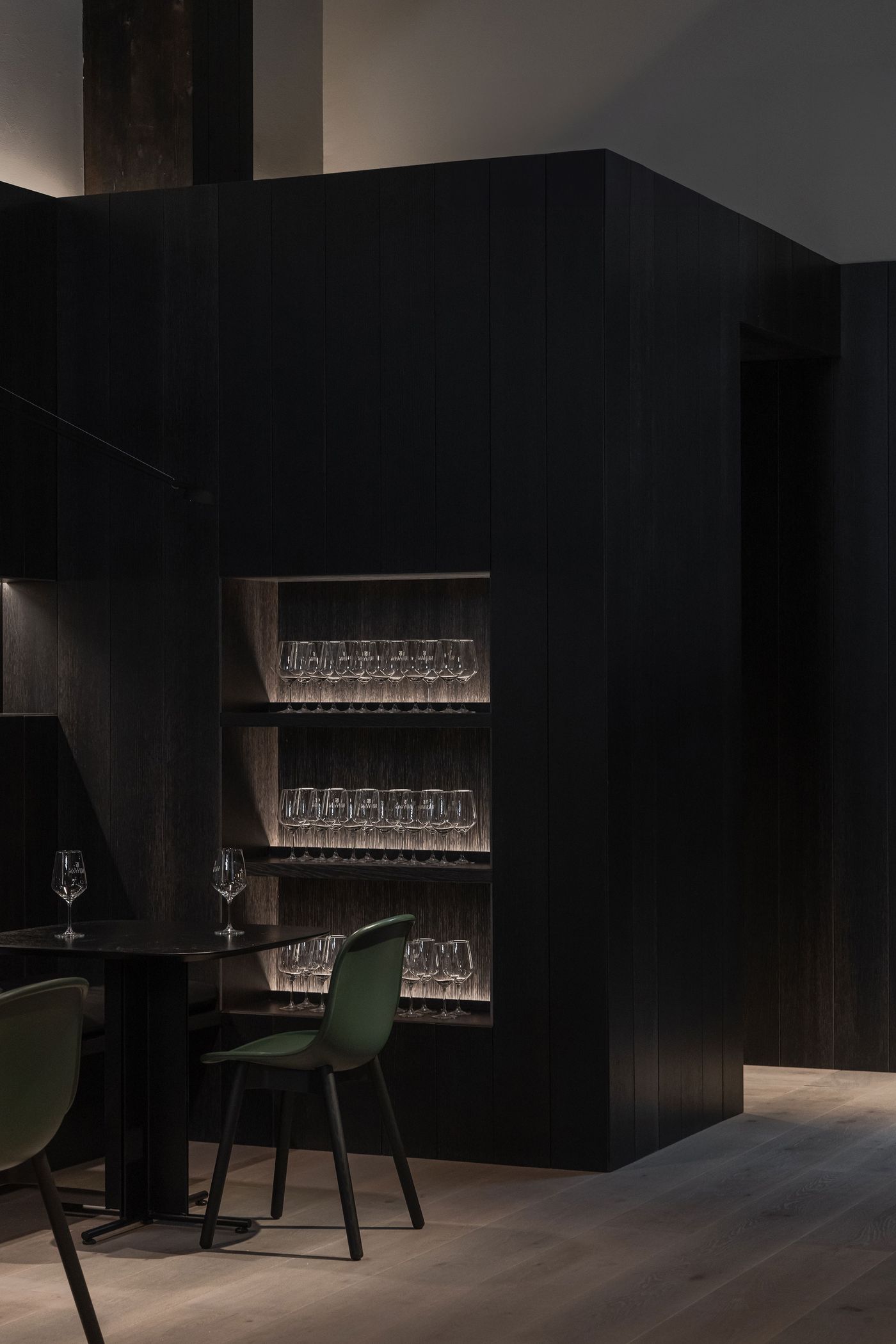A dark, stylish restaurant interior featuring a sleek table, green chair, and illuminated shelves lined with wine glasses.