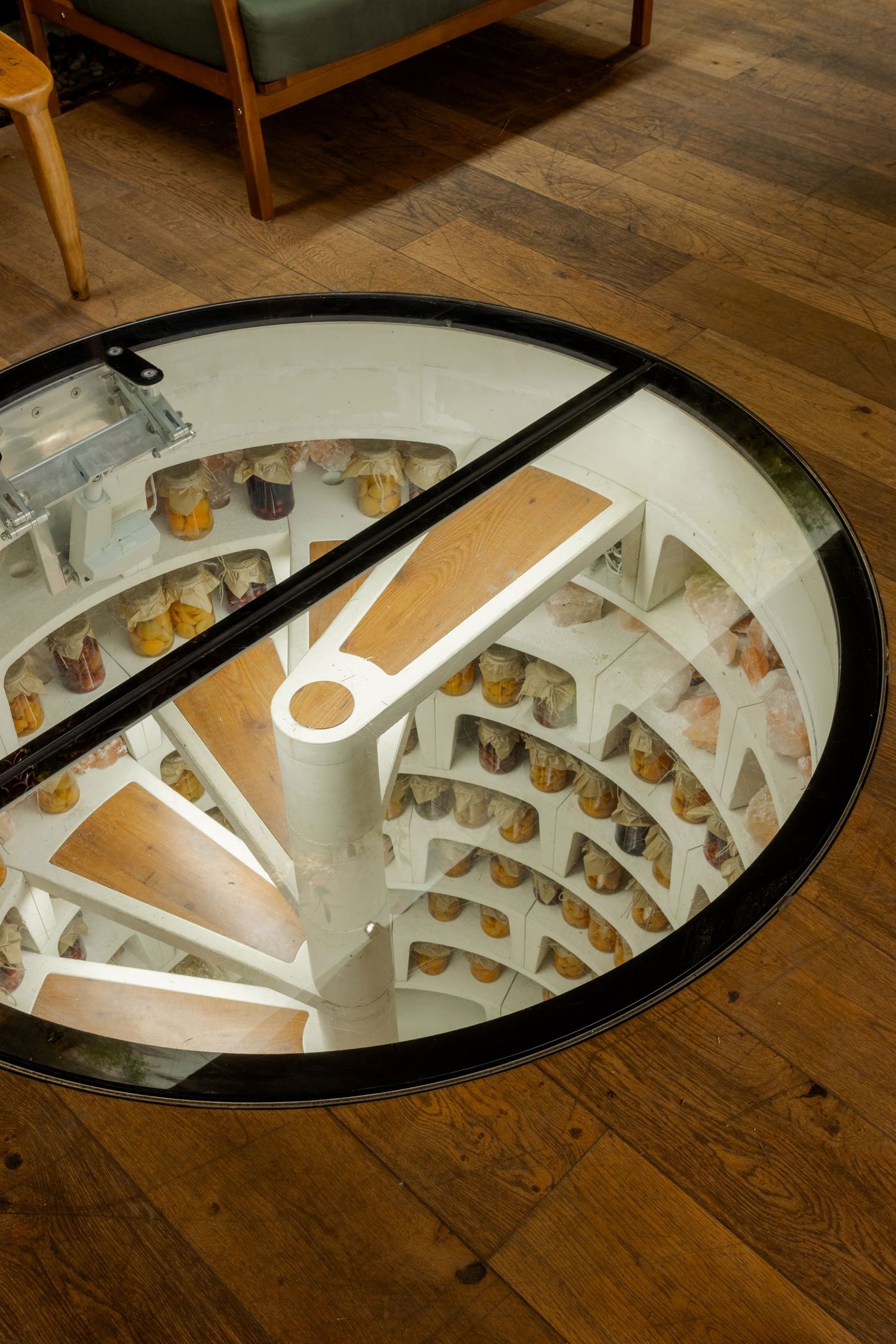 Circular glass opening on the floor, revealing a spiral staircase and jars on shelves below, surrounded by wooden flooring and furniture above.