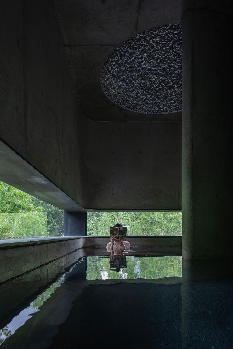 A person immersed in water, holding a book, surrounded by minimalist concreA person showers beneath a circular concrete structure, with water cascading down from above, illuminated by natural light.te architecture and green trees visible through large windows. A person immersed in water, holding a book, surrounded by minimalist concreA person showers beneath a circular concrete structure, with water cascading down from above, illuminated by natural light.te architecture and green trees visible through large windows.