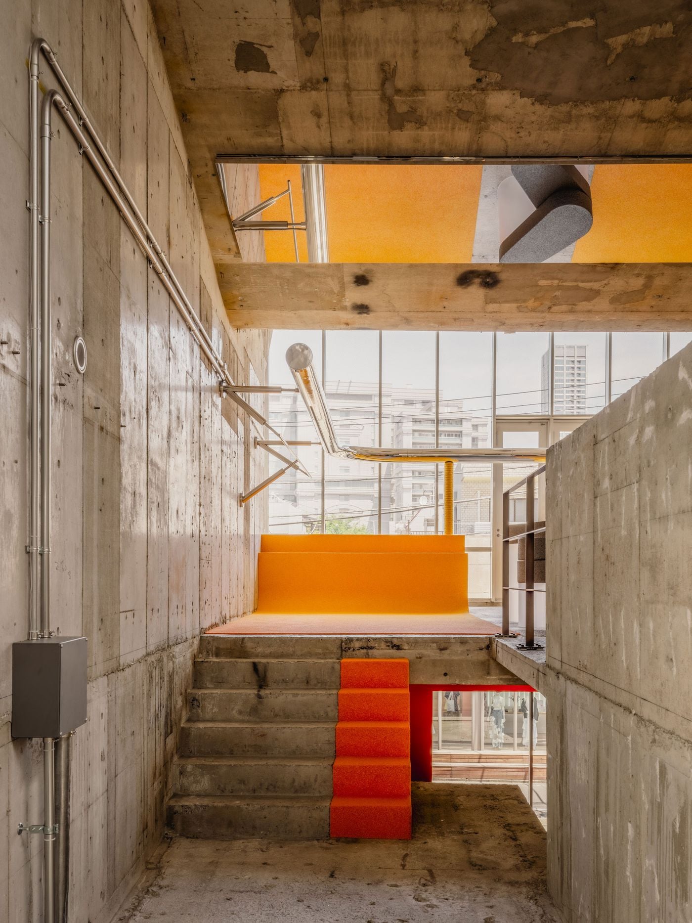 A raw concrete stair rises towards the yellow upper level, punctuated by a narrow strip of orange steps that reads almost like a freestanding object. Above, a large ceiling opening offers glimpses of the floor beyond, reinforcing the store’s deliberately porous, vertically interconnected composition.