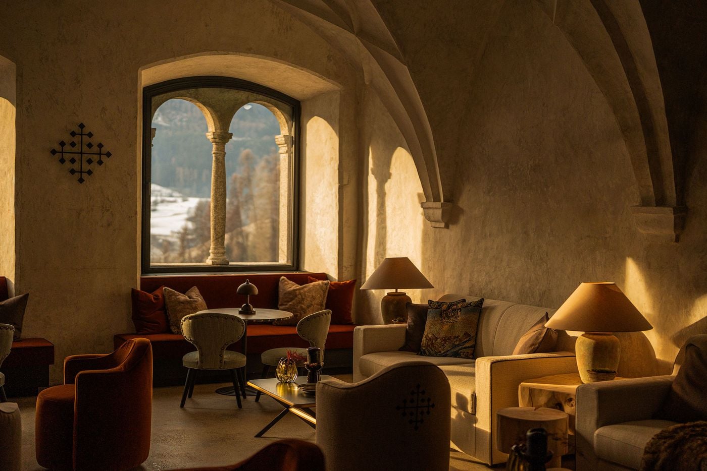 Low winter sun pours through a twin-arched stone window into the vaulted former Abbess's Hall at Castel Badia, casting the column's shadow across lime-washed walls. A rust velvet banquette, studded chairs, cream sofas, terracotta lamps and a wrought-iron cross emblem furnish the lounge. Low winter sun pours through a twin-arched stone window into the vaulted former Abbess's Hall at Castel Badia, casting the column's shadow across lime-washed walls. A rust velvet banquette, studded chairs, cream sofas, terracotta lamps and a wrought-iron cross emblem furnish the lounge.