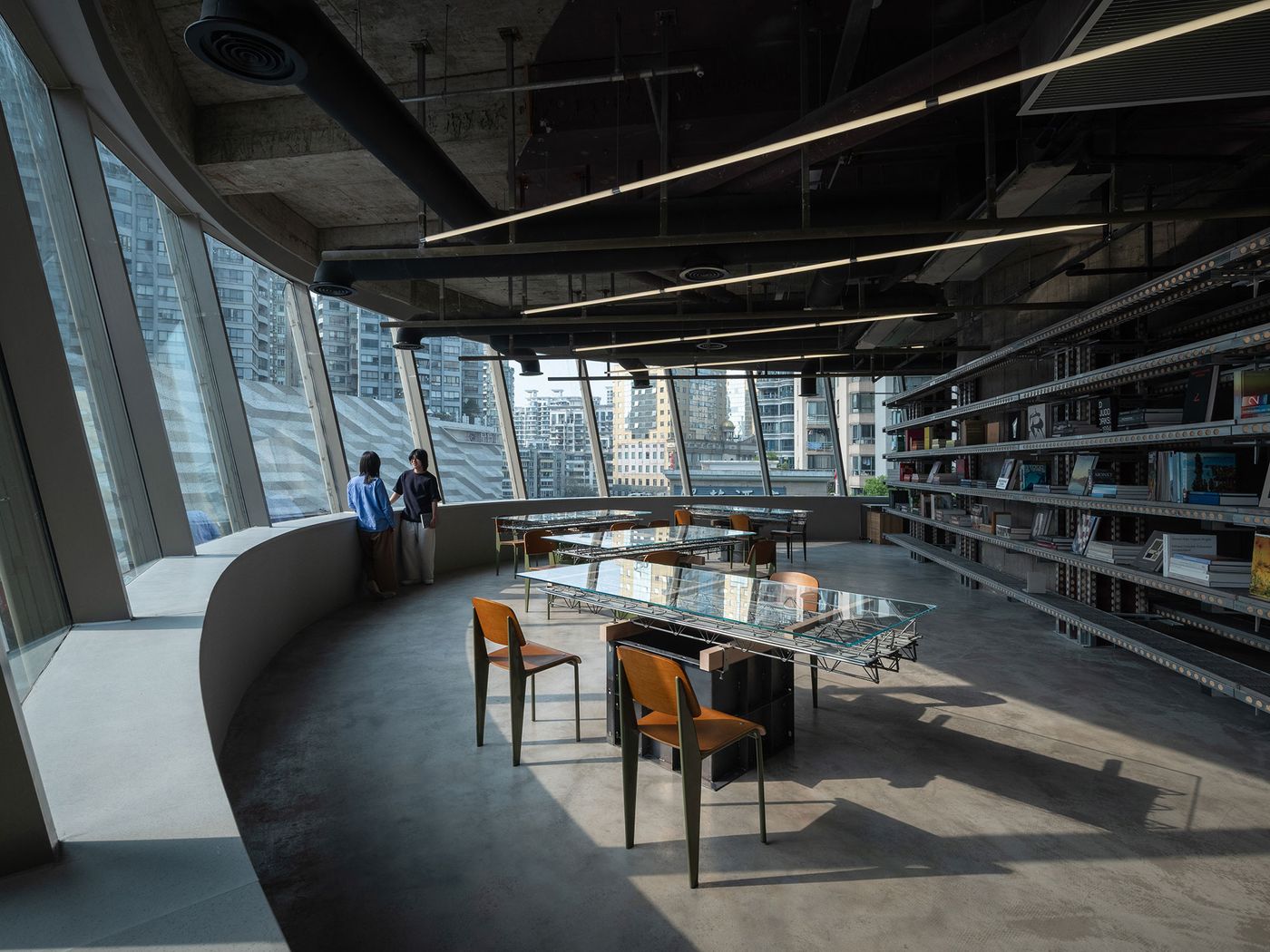 A sweeping view of 889 GLO Art Space's reading area, offering panoramic city views through expansive, curved windows. The minimalist concrete floor is complemented by glass-topped tables with industrial bases and modern chairs. A vast, industrial shelving unit filled with books stretches along one wall, balancing the raw architecture with a sophisticated, intellectual atmosphere. A sweeping view of 889 GLO Art Space's reading area, offering panoramic city views through expansive, curved windows. The minimalist concrete floor is complemented by glass-topped tables with industrial bases and modern chairs. A vast, industrial shelving unit filled with books stretches along one wall, balancing the raw architecture with a sophisticated, intellectual atmosphere.