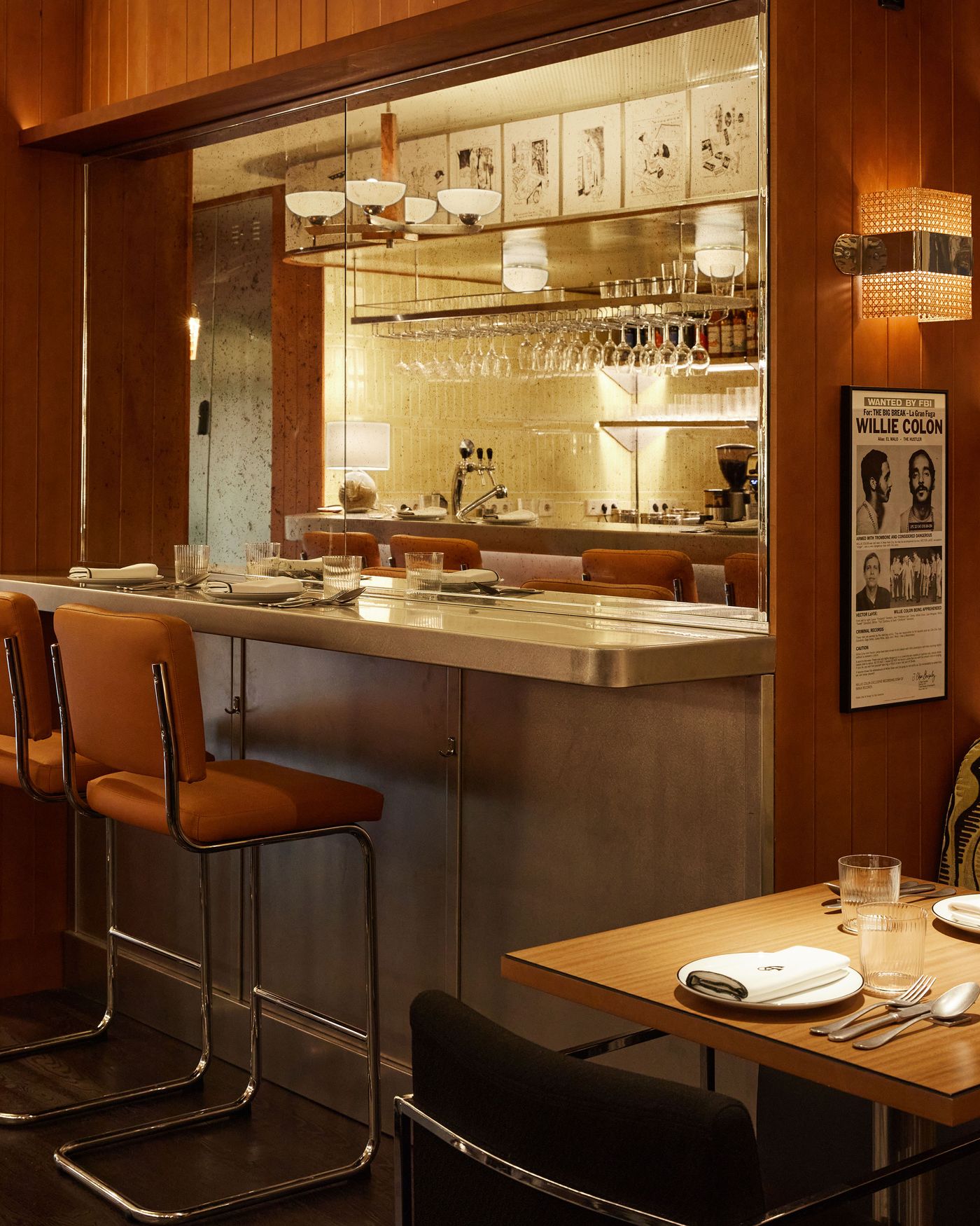 This interior view highlights a sophisticated retro-industrial aesthetic. The stainless steel bar counter and bar stools with tan leather seats create a sleek, classic look. A large mirror reflects the bar area, while framed art and a textured woven sconce add warmth and artistic detail. This interior view highlights a sophisticated retro-industrial aesthetic. The stainless steel bar counter and bar stools with tan leather seats create a sleek, classic look. A large mirror reflects the bar area, while framed art and a textured woven sconce add warmth and artistic detail.