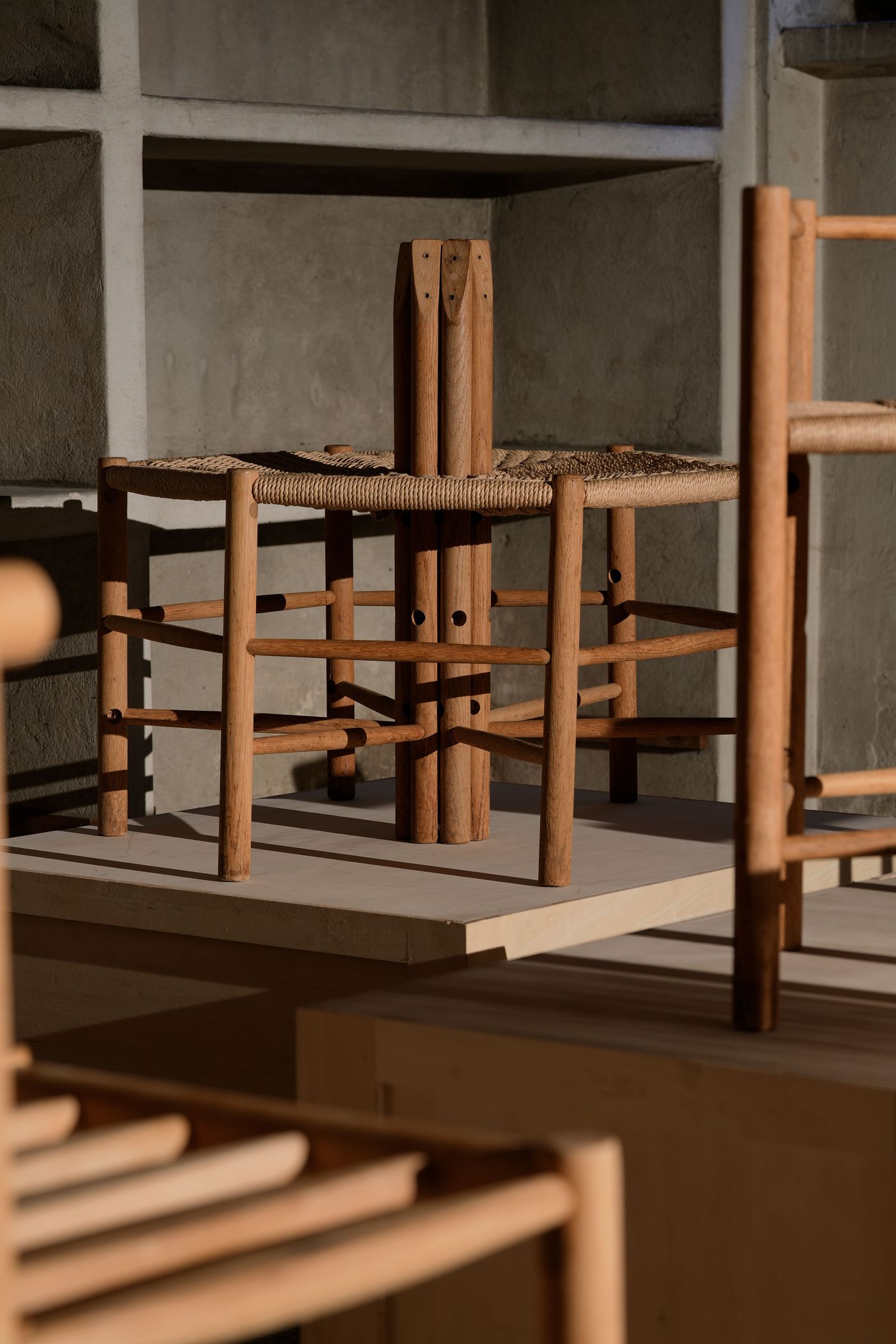 A collection of wooden chairs with woven seats displayed on a light-colored platform against a textured wall. Soft shadows enhance features. A collection of wooden chairs with woven seats displayed on a light-colored platform against a textured wall. Soft shadows enhance features.