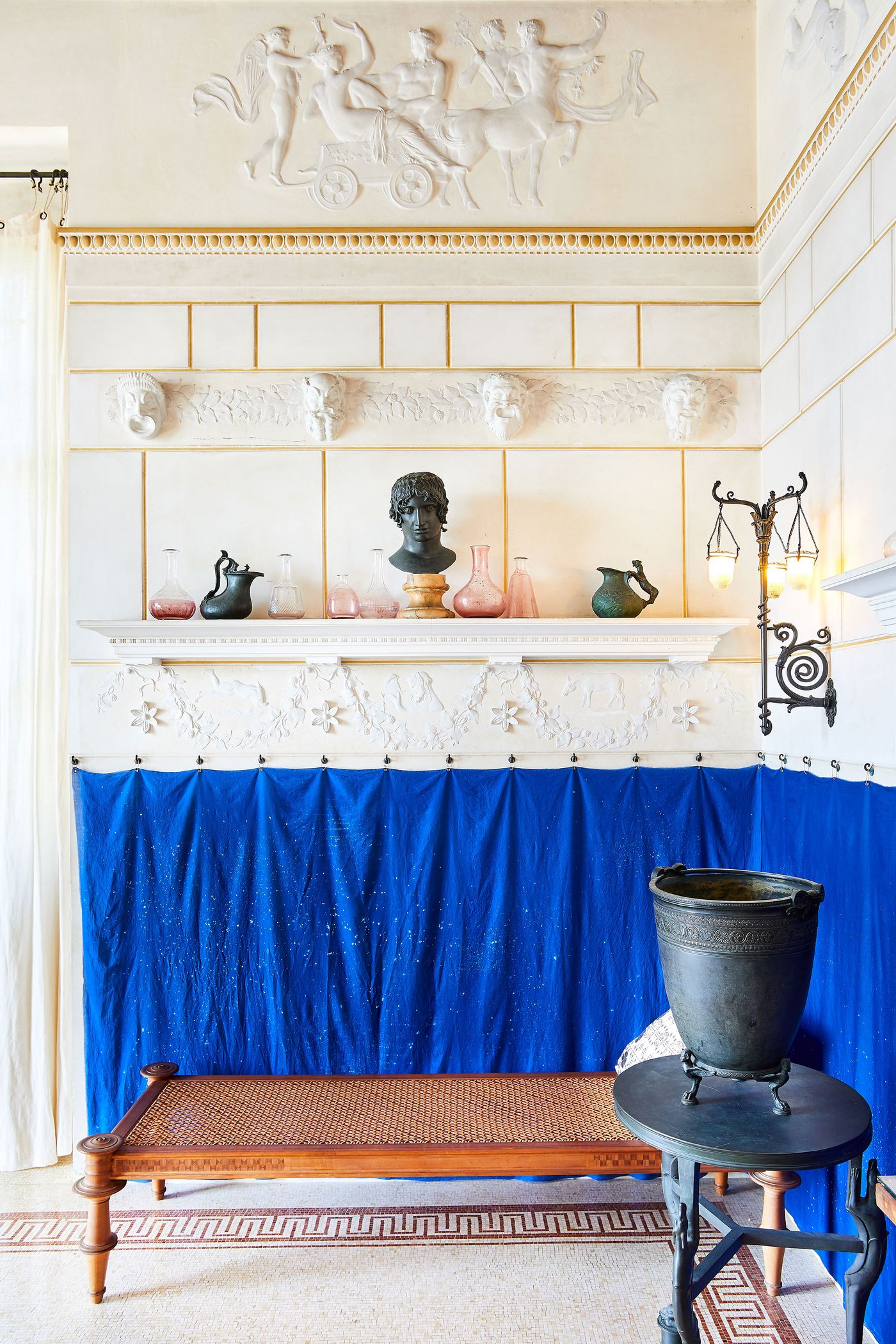 A Grecian-styled room with a half wall of draped cobalt blue curtain, contrasting with the pale yellow walls above. These walls are decorated with white plaster friezes and a central shelf displaying classical bronze and colored glass vessels. A woven chaise lounge and a rustic stool with a metal basin sit on a mosaic floor, balancing the room's elements. A Grecian-styled room with a half wall of draped cobalt blue curtain, contrasting with the pale yellow walls above. These walls are decorated with white plaster friezes and a central shelf displaying classical bronze and colored glass vessels. A woven chaise lounge and a rustic stool with a metal basin sit on a mosaic floor, balancing the room's elements.