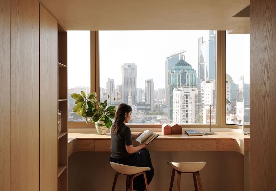 A woman engrossed in a book at a beautifully integrated wooden desk, framed by an expansive window overlooking the vibrant Shanghai cityscape. The minimalist workstation is part of a custom wooden built-in that includes shelving, creating a serene and productive environment that harmoniously connects with the urban panorama.