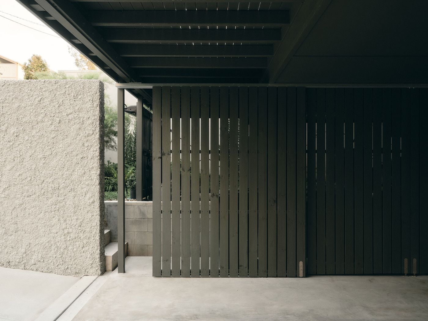 Modern architectural entrance featuring a textured gray wall and a dark wooden slatted gate under a shaded overhang. Modern architectural entrance featuring a textured gray wall and a dark wooden slatted gate under a shaded overhang.