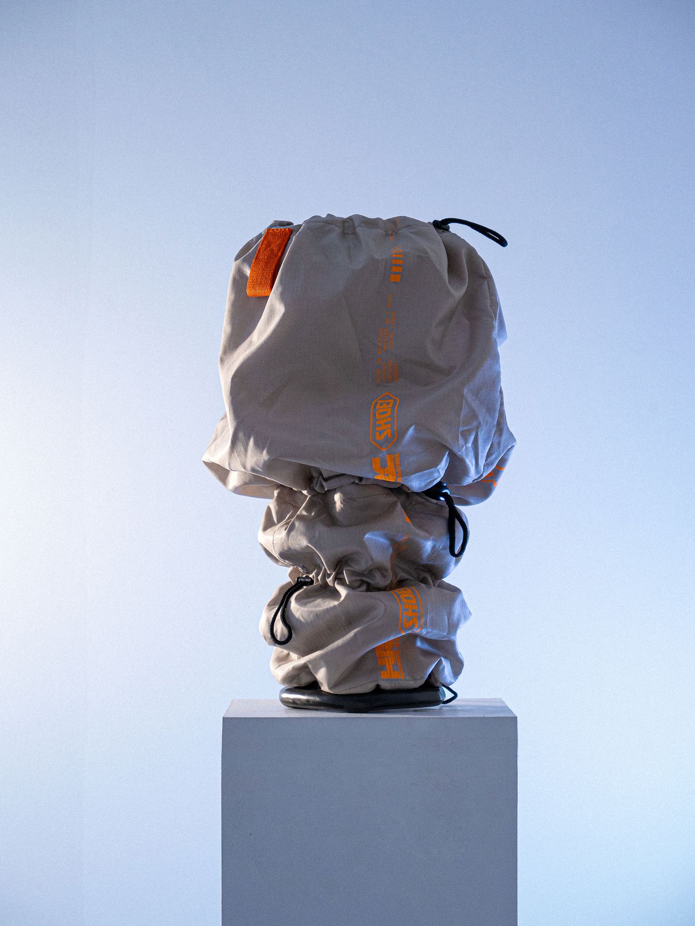 A beige bag with an orange logo, cinched at the top, sits on a pedestal against a light blue background, showcasing its unique design.