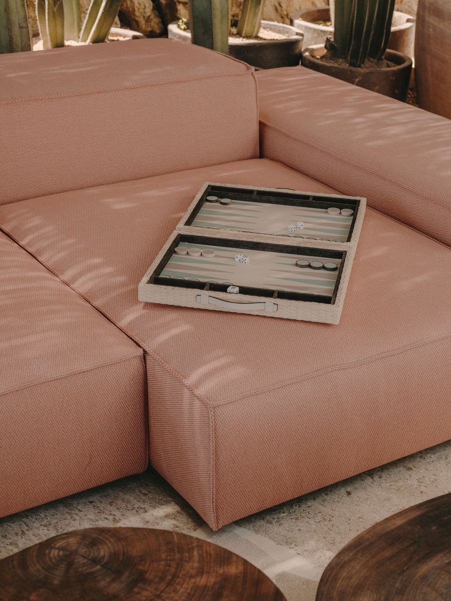 A close-up of a pink sectional sofa with a backgammon set resting on it, surrounded by cacti and wooden tables in a sunny outdoor setting. A close-up of a pink sectional sofa with a backgammon set resting on it, surrounded by cacti and wooden tables in a sunny outdoor setting.
