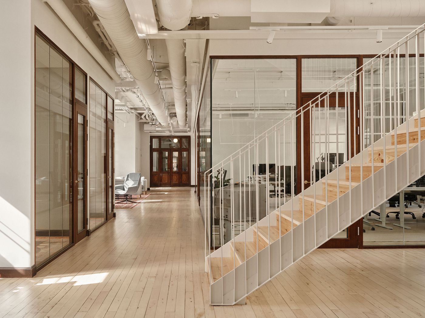 A bright industrial office corridor at Konelabra, Helsinki by Fyra, characterized by light wooden floors and exposed white ceiling pipes. Glass-walled offices with dark wood frames line one side, leading to a distant seating area. A prominent white metal staircase with light wood treads ascends, embodying a blend of raw industrial aesthetics and refined Scandinavian design. A bright industrial office corridor at Konelabra, Helsinki by Fyra, characterized by light wooden floors and exposed white ceiling pipes. Glass-walled offices with dark wood frames line one side, leading to a distant seating area. A prominent white metal staircase with light wood treads ascends, embodying a blend of raw industrial aesthetics and refined Scandinavian design.