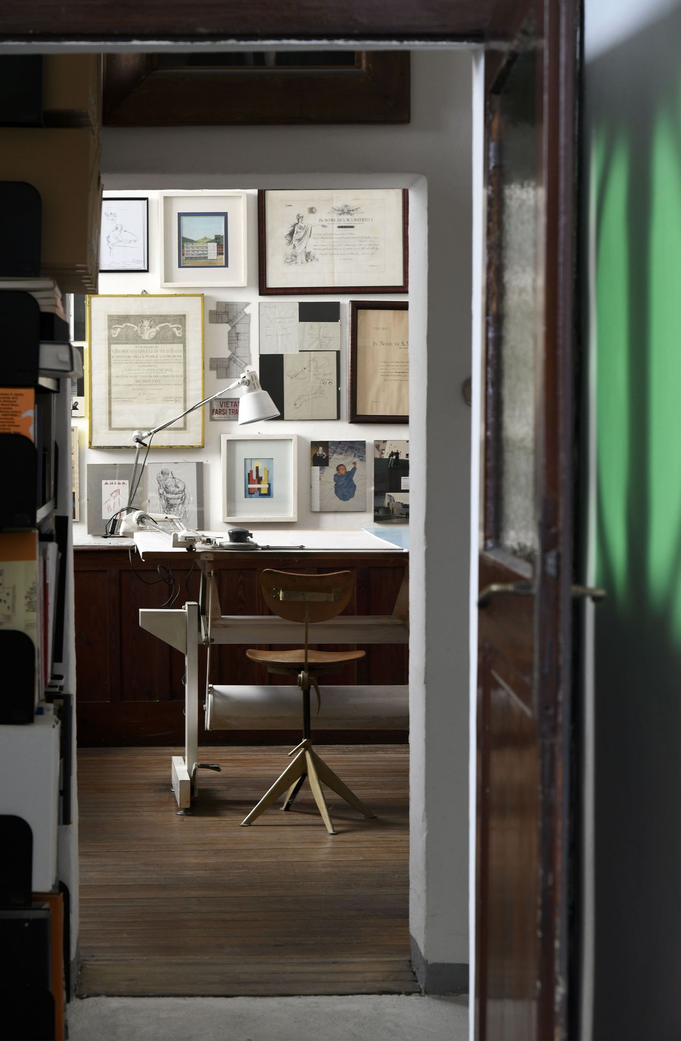 A brightly lit workspace is viewed through an open doorway, revealing a desk with a vintage lamp and a simple wooden stool. The wall behind is covered with an eclectic arrangement of framed documents, artworks, and photographs. A large bookcase is partially visible to the left. The scene conveys a bohemian and scholarly atmosphere, with a focus on art, literature, and design.