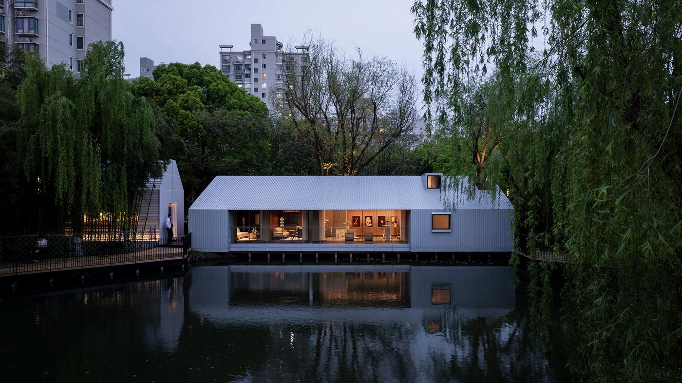 Dusk settles over Wutopia Lab's Lake House, its gabled, corrugated metal form reflecting the fading light. Warm illumination from within reveals an art-filled interior, creating an inviting glow across the lake. The building, framed by dark weeping willows, acts as a serene counterpoint to the distant, softly lit urban structures, embodying a peaceful modern retreat. Dusk settles over Wutopia Lab's Lake House, its gabled, corrugated metal form reflecting the fading light. Warm illumination from within reveals an art-filled interior, creating an inviting glow across the lake. The building, framed by dark weeping willows, acts as a serene counterpoint to the distant, softly lit urban structures, embodying a peaceful modern retreat.