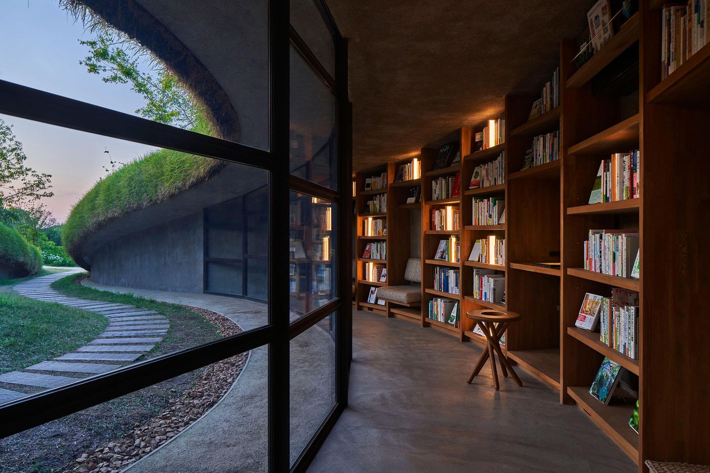 A cozy library with wooden shelves full of books, featuring a view of a curved path and grassy rooftop through glass doors. A cozy library with wooden shelves full of books, featuring a view of a curved path and grassy rooftop through glass doors.