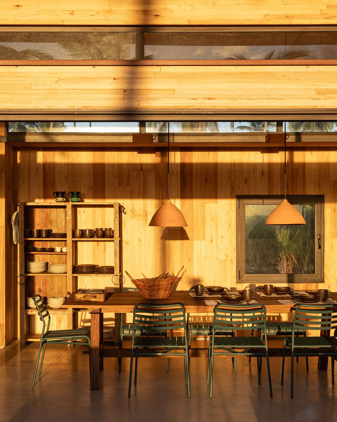 A warm, timber-clad dining area bathed in golden light features a long rustic table, modern green chairs, and open shelving filled with ceramics. Ceramic pendant lights and horizontal windows revealing glimpses of palm trees enhance the inviting, natural aesthetic. A warm, timber-clad dining area bathed in golden light features a long rustic table, modern green chairs, and open shelving filled with ceramics. Ceramic pendant lights and horizontal windows revealing glimpses of palm trees enhance the inviting, natural aesthetic.