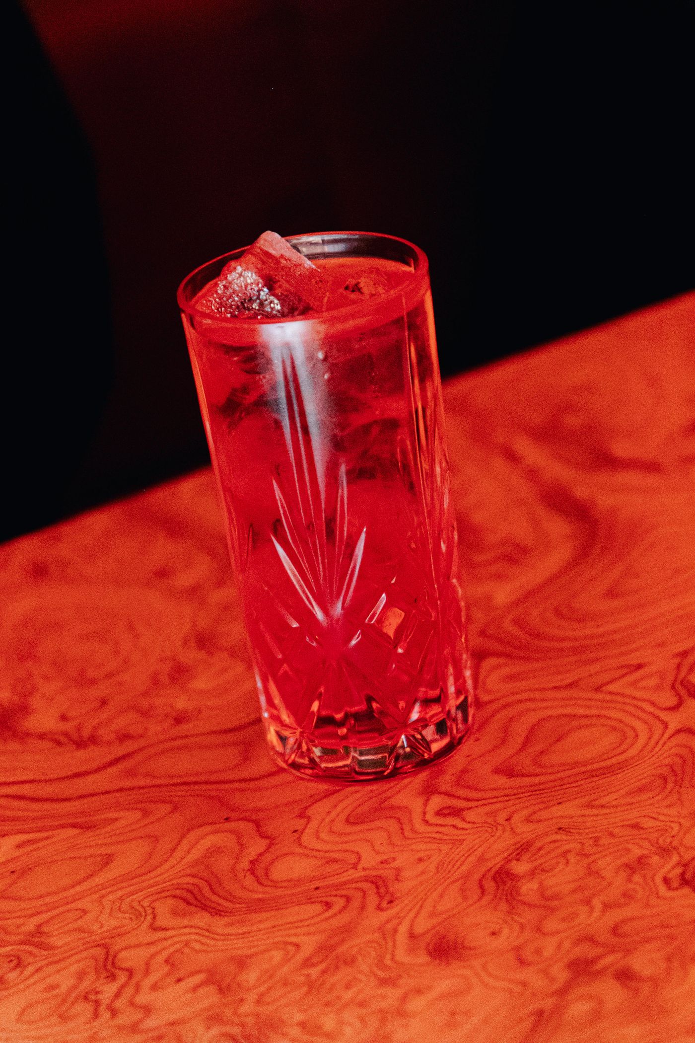 A crystal glass filled with a vibrant red drink and ice, sitting on a smooth, patterned wooden table.