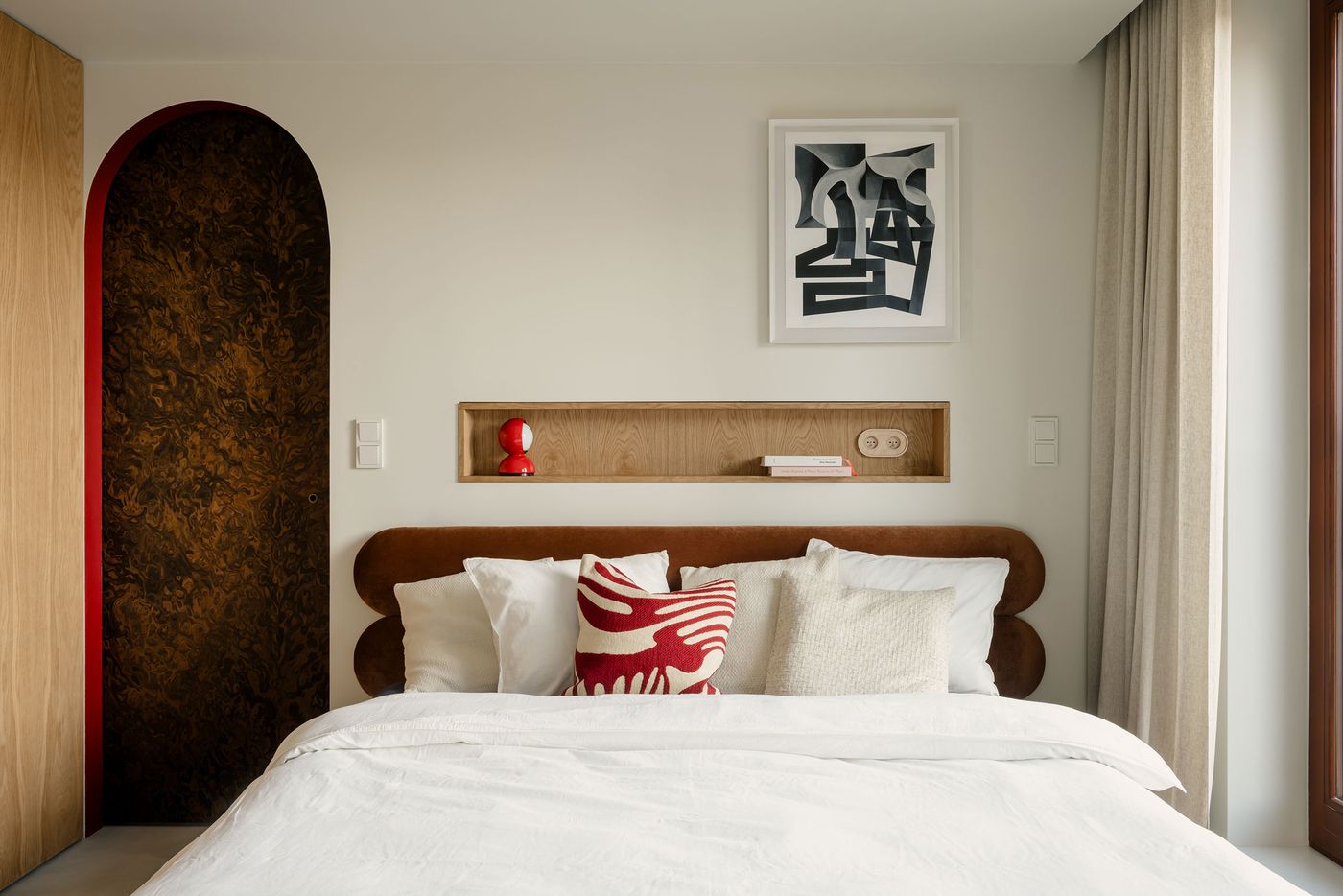 A calm, light-filled bedroom blends neutral tones with warm wood and a soft suede headboard. A pop of red animates the composition—from a sculptural lamp and a graphic cushion to the deep red trim of the curved doorframe. A calm, light-filled bedroom blends neutral tones with warm wood and a soft suede headboard. A pop of red animates the composition—from a sculptural lamp and a graphic cushion to the deep red trim of the curved doorframe.