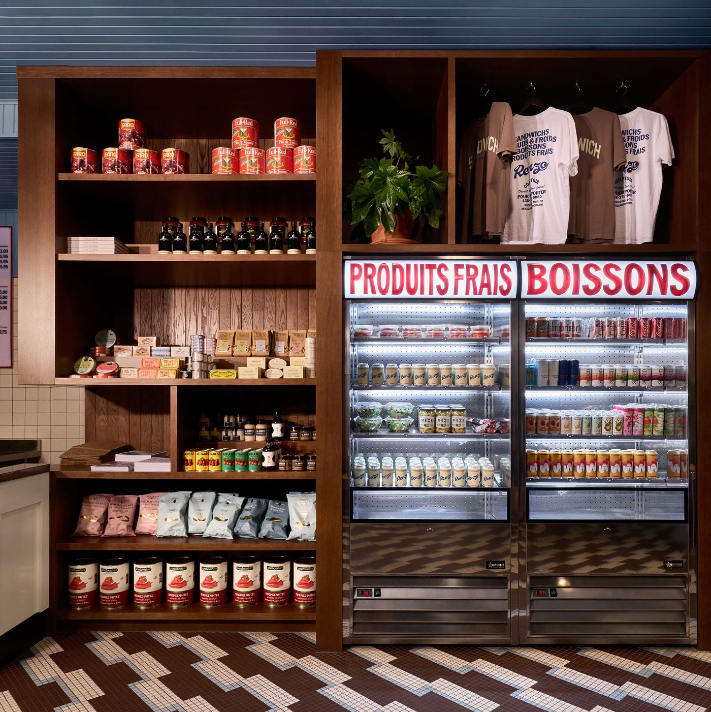 This image showcases the retail display at Renzo Sandwich Shop, featuring wooden shelves stocked with various canned goods, sauces, and packaged snacks. Two large, illuminated refrigerators are filled with beverages. T-shirts with the shop's branding hang above the coolers, emphasizing the brand's presence and offerings.