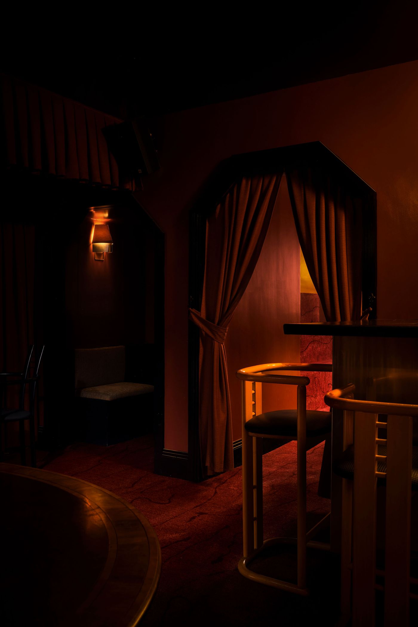 A moody, inviting nook at The Roses of Elagabalus, characterized by deep red walls and a curved, dark wooden bar section with sleek chairs. An arched doorway, framed by a tied-back red curtain, reveals a glimpse of another warmly lit space beyond, contributing to the establishment's secretive and alluring vintage-retro atmosphere. A moody, inviting nook at The Roses of Elagabalus, characterized by deep red walls and a curved, dark wooden bar section with sleek chairs. An arched doorway, framed by a tied-back red curtain, reveals a glimpse of another warmly lit space beyond, contributing to the establishment's secretive and alluring vintage-retro atmosphere.