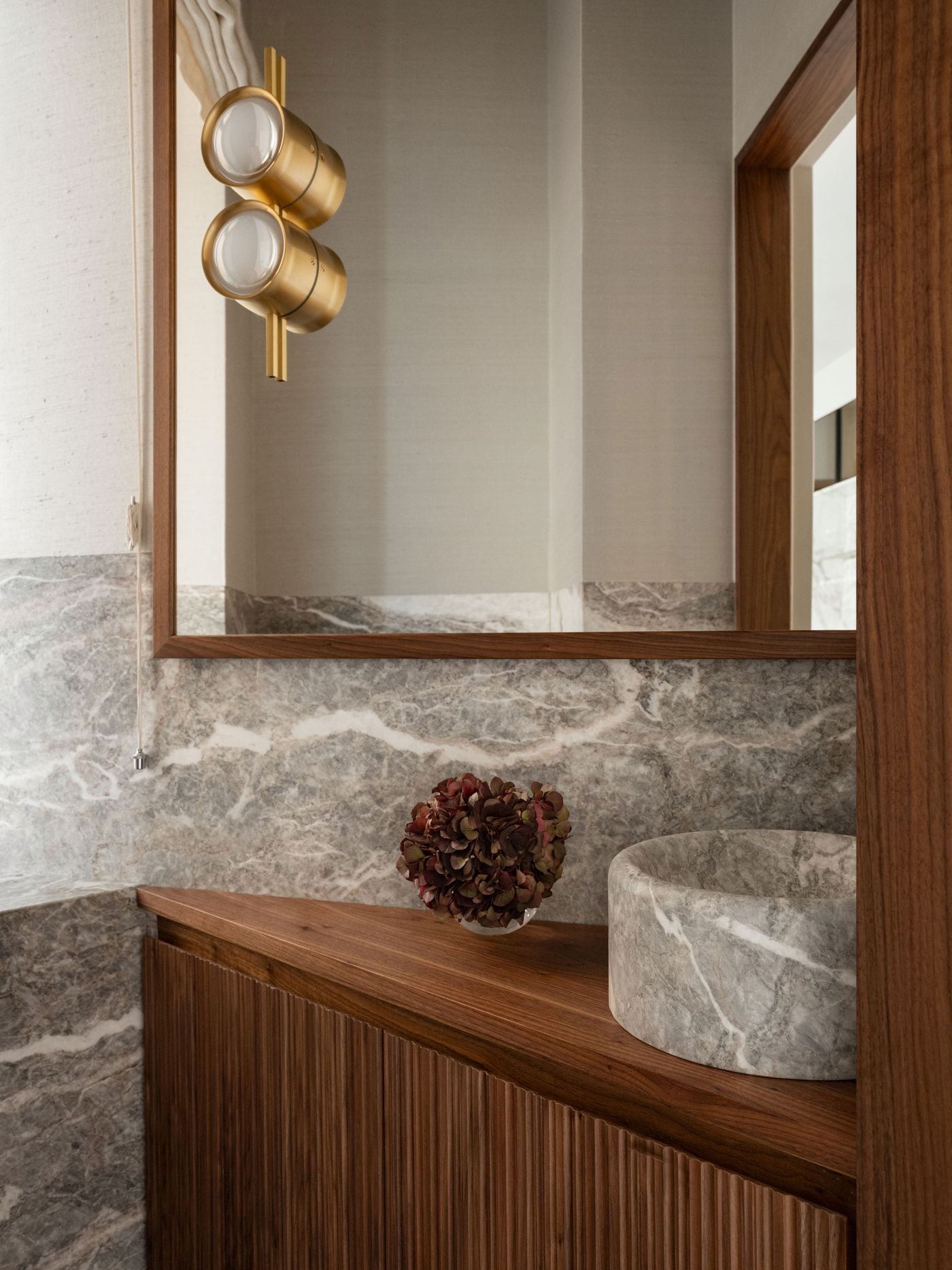 A modern bathroom features a marble sink, a wooden countertop, a decorative floral arrangement, and stylish gold light fixtures.