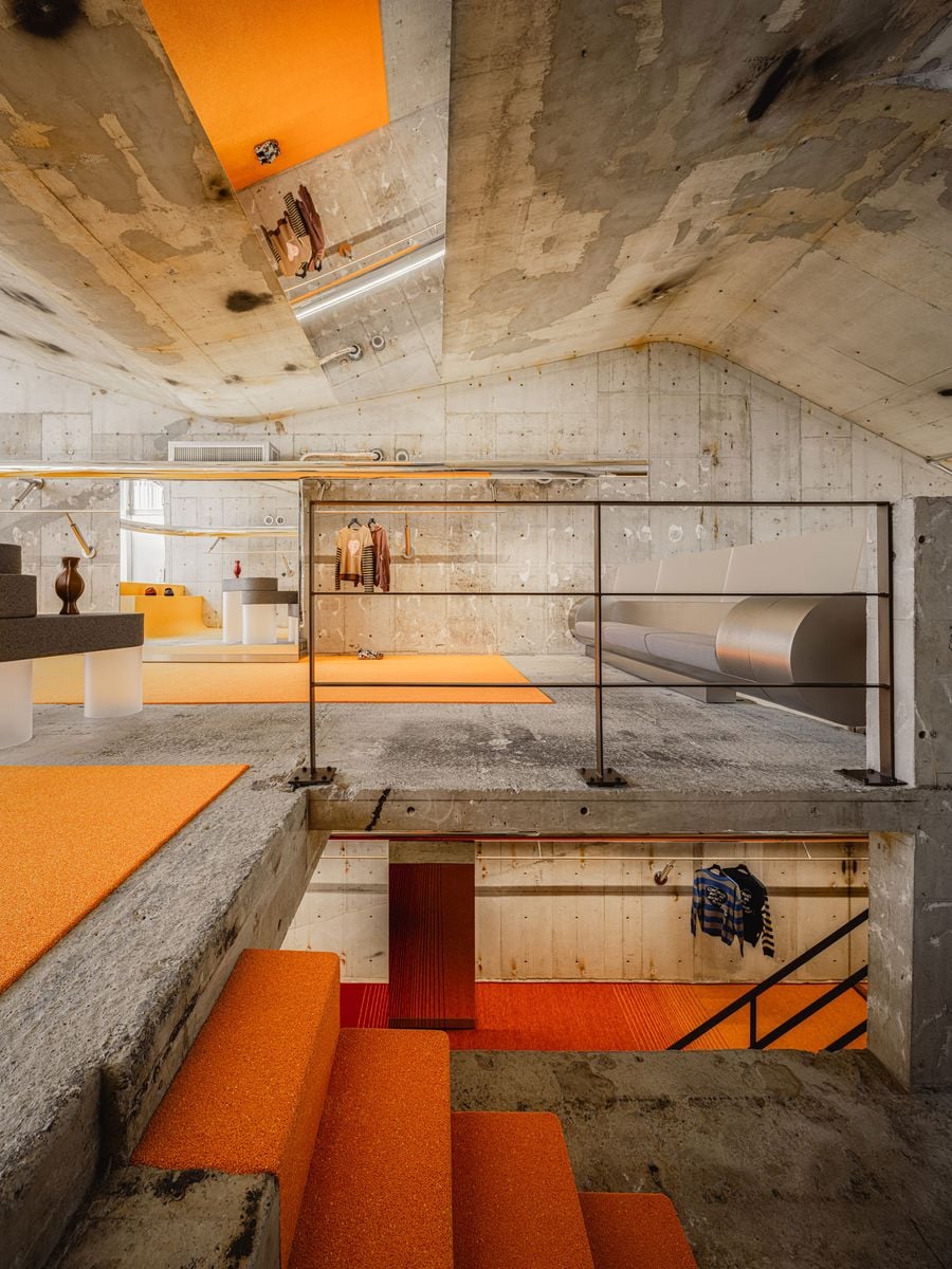 A broad opening reveals the store’s layered section, with orange-covered steps descending towards the ground floor while the upper level stretches beyond a slender black balustrade. Exposed slabs, patched concrete and mirrored surfaces preserve long sightlines, allowing different colours, displays and levels to register simultaneously.