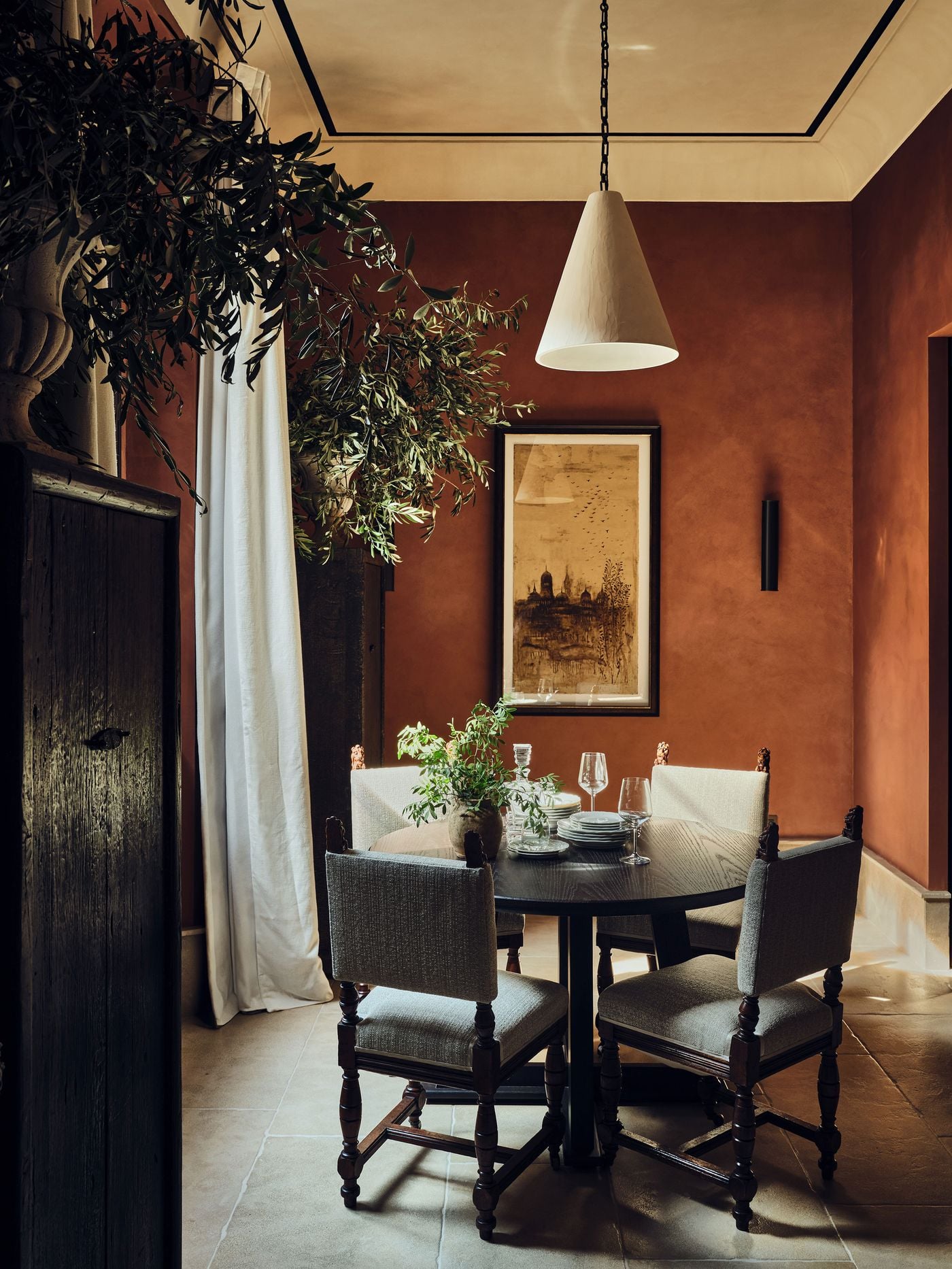 Seen from another angle, the terracotta-toned dining room at La Réserve Firenze reveals a framed work of a domed city skyline scattered with birds. A conical plaster pendant, a slim black wall light and antique lion-finial chairs around a round table bring warmth and intimacy, framed by olive branches and a weathered cabinet. Seen from another angle, the terracotta-toned dining room at La Réserve Firenze reveals a framed work of a domed city skyline scattered with birds. A conical plaster pendant, a slim black wall light and antique lion-finial chairs around a round table bring warmth and intimacy, framed by olive branches and a weathered cabinet.