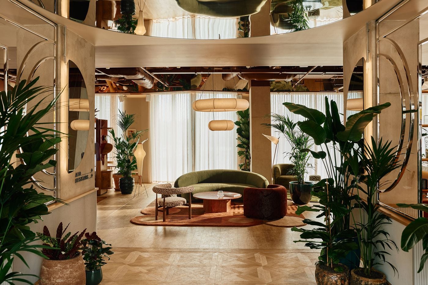 Viewed from the entrance, the lounge unfolds as a lush, theatrical composition framed by mirrored panels and abundant planting. Curved olive seating, patterned rugs and warm timber sit beneath sculptural pendant lights, while the reflections overhead amplify Holloway Li’s mix of biophilic softness and mid-century-inspired glamour. Viewed from the entrance, the lounge unfolds as a lush, theatrical composition framed by mirrored panels and abundant planting. Curved olive seating, patterned rugs and warm timber sit beneath sculptural pendant lights, while the reflections overhead amplify Holloway Li’s mix of biophilic softness and mid-century-inspired glamour.