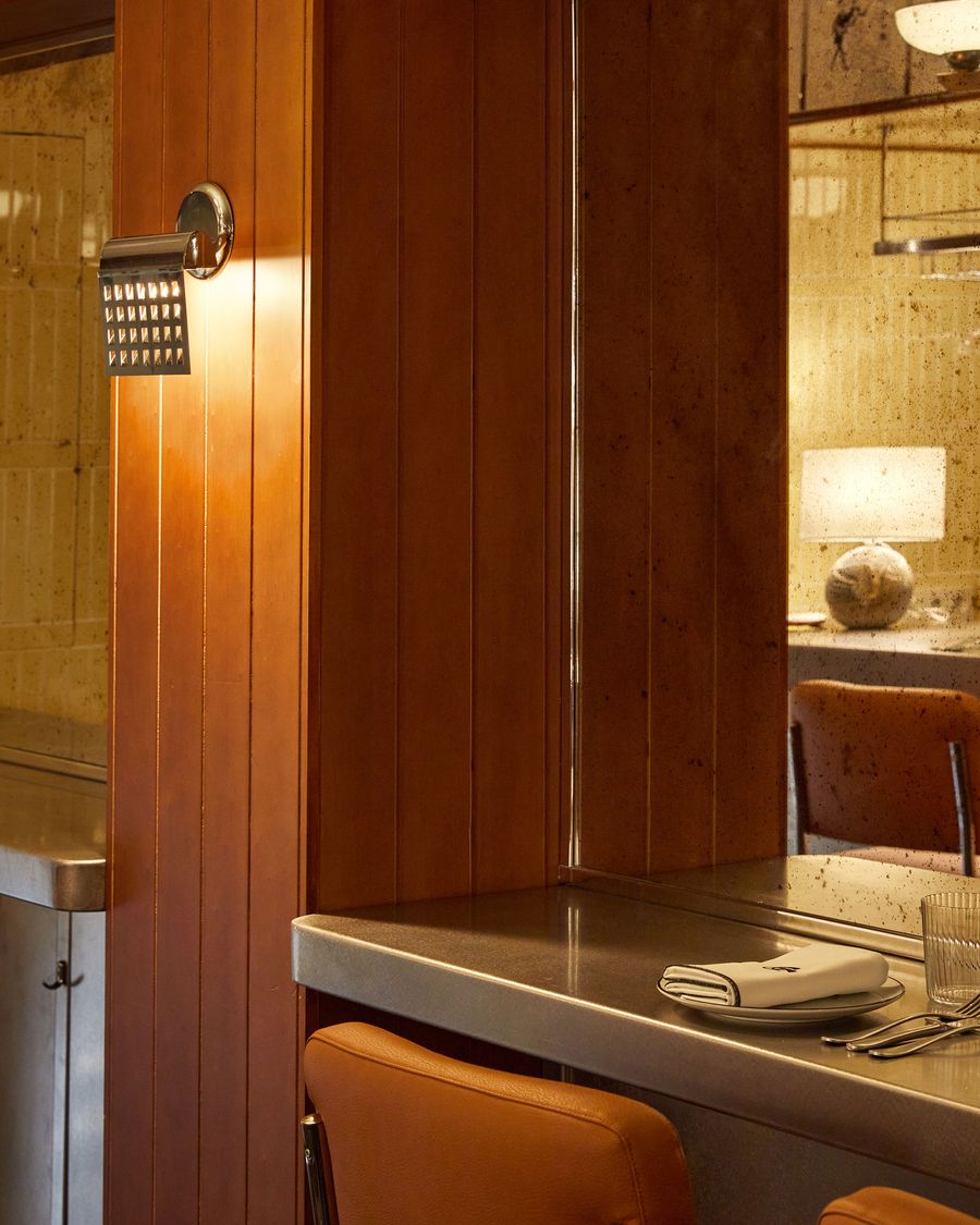 A detail shot focusing on the interplay of light and materials. A vintage-inspired metal sconce with a perforated screen casts a warm light on a rich, wood-paneled wall. The reflection in the adjacent mottled mirror reveals a lamp and bar stools, adding depth and a sense of inviting atmosphere. A detail shot focusing on the interplay of light and materials. A vintage-inspired metal sconce with a perforated screen casts a warm light on a rich, wood-paneled wall. The reflection in the adjacent mottled mirror reveals a lamp and bar stools, adding depth and a sense of inviting atmosphere.