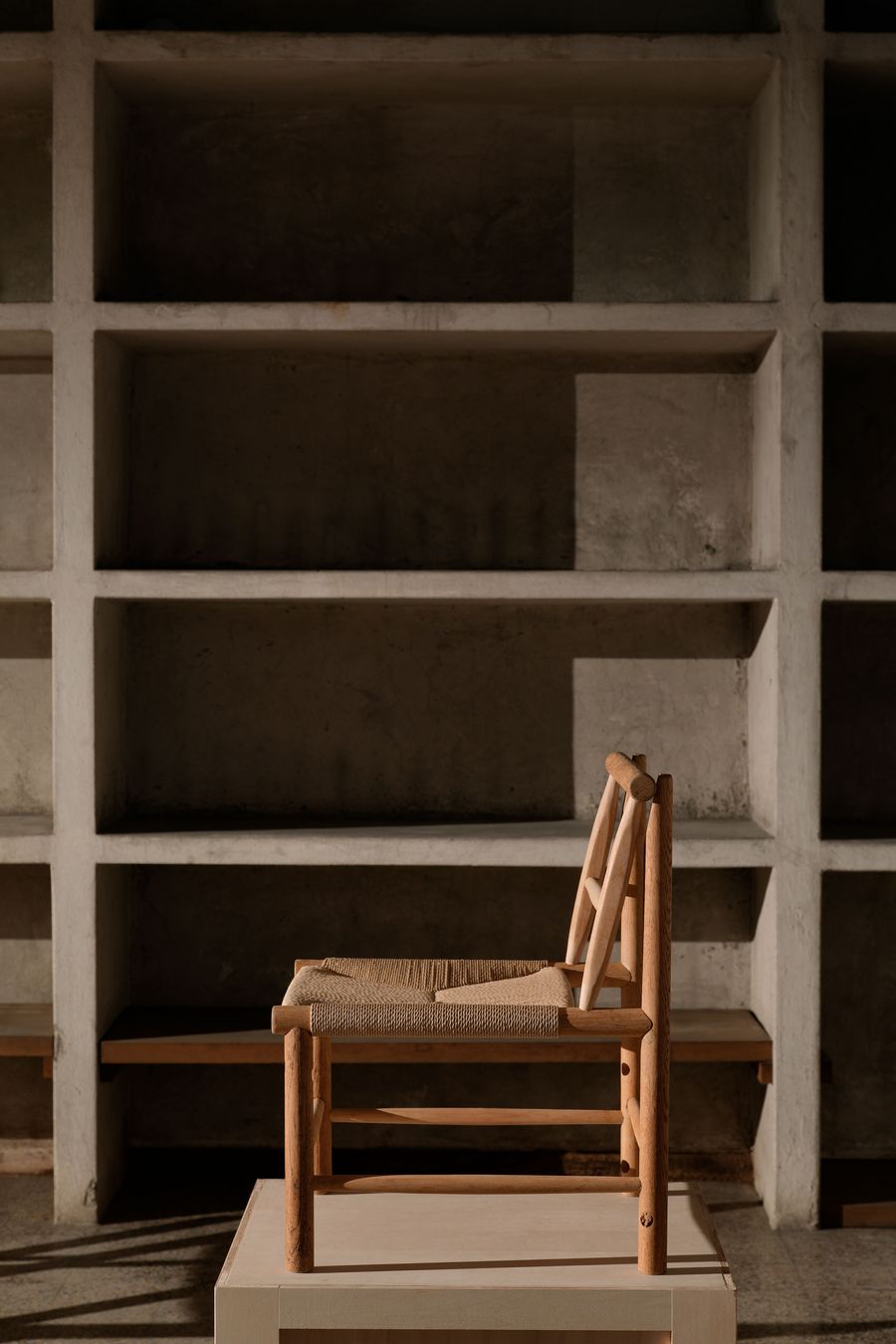 A simple wooden chair with a woven seat positioned on a platform against a minimalistic, concrete shelf background. A simple wooden chair with a woven seat positioned on a platform against a minimalistic, concrete shelf background.