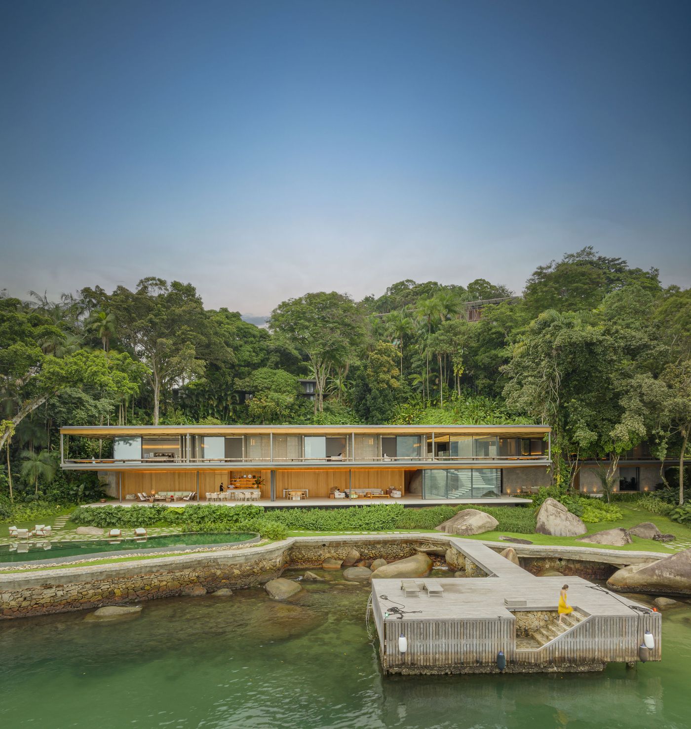 The expansive horizontal residence stands gracefully at the water's edge, its warm wooden facades and extensive glazing glowing with interior light against the twilight sky. A series of natural pools and a timber dock lead into the calm waters, showcasing a harmonious blend of architecture and a vibrant coastal landscape. The expansive horizontal residence stands gracefully at the water's edge, its warm wooden facades and extensive glazing glowing with interior light against the twilight sky. A series of natural pools and a timber dock lead into the calm waters, showcasing a harmonious blend of architecture and a vibrant coastal landscape.
