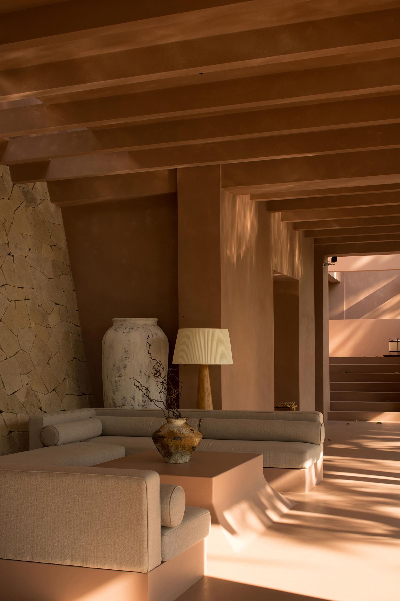 A modern living space with warm brown tones, featuring a sectional sofa, decorative vase, lamp, and a textured stone wall. A modern living space with warm brown tones, featuring a sectional sofa, decorative vase, lamp, and a textured stone wall.