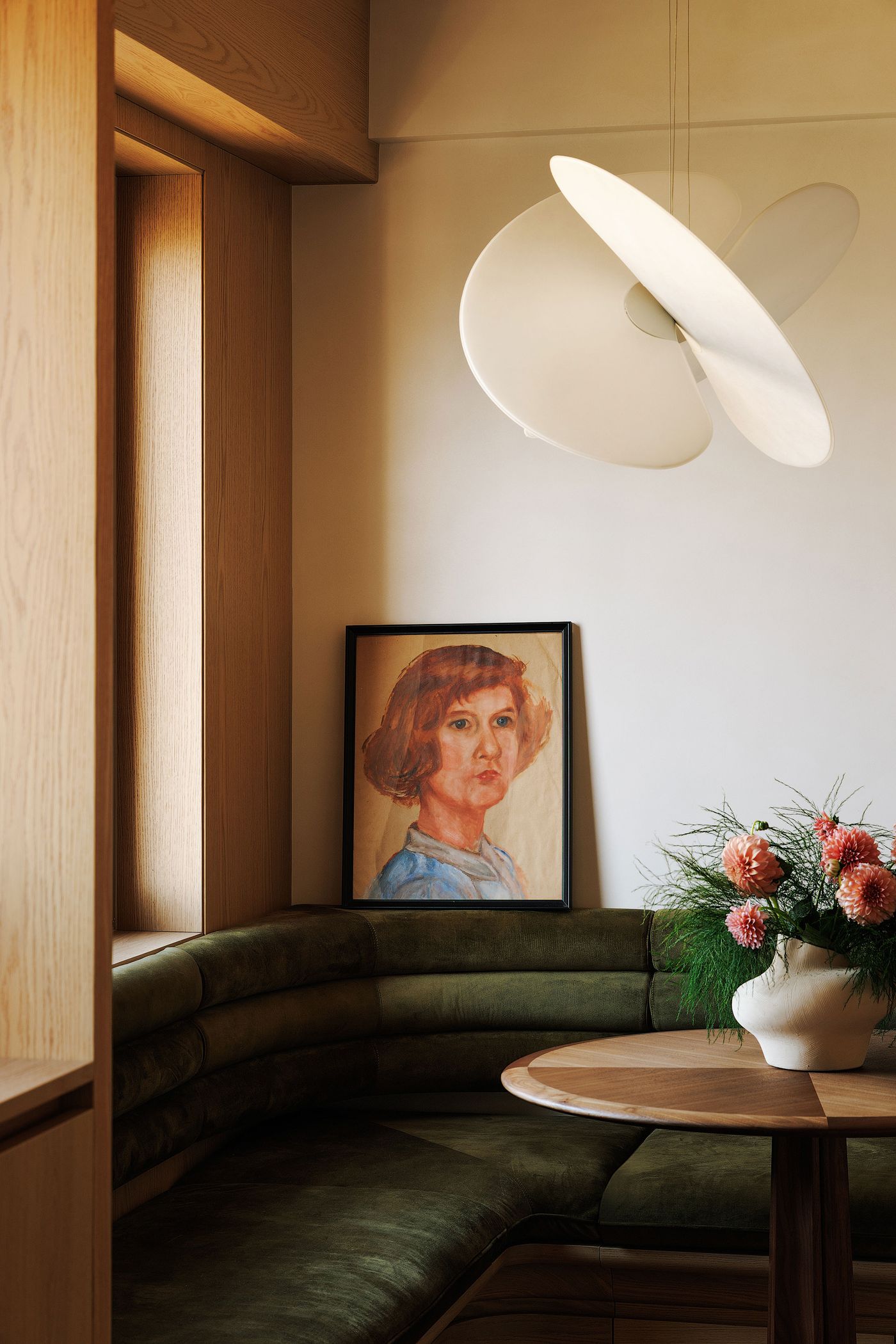 A charming dining nook featuring an L-shaped banquette upholstered in rich olive green velvet, set against warm wooden walls. A round wooden table with a unique two-tone top is adorned with a vibrant floral arrangement. A classical portrait hangs above the banquette, while a sculptural white pendant light casts an elegant glow, creating an intimate and artful atmosphere.