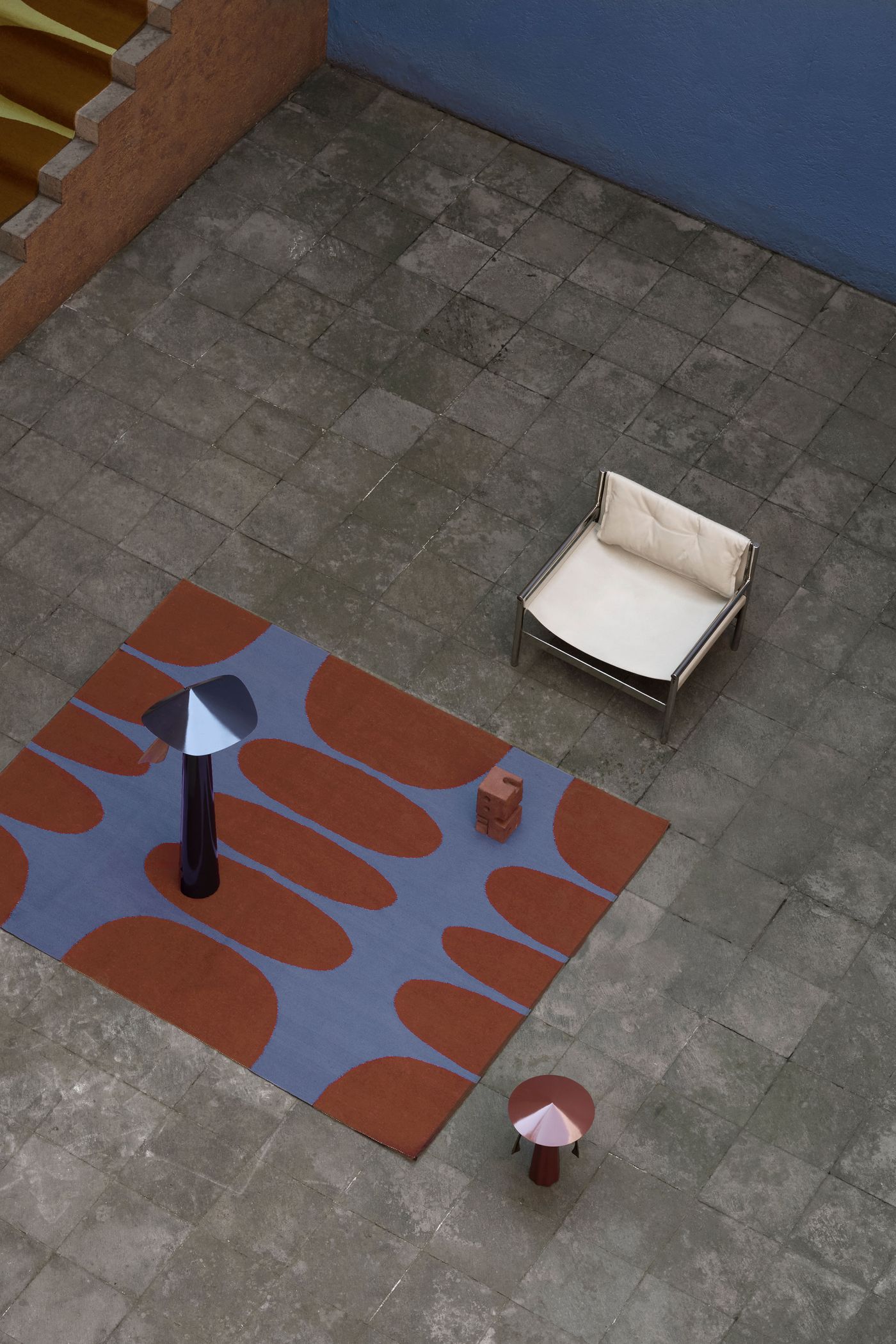 A modern outdoor scene with a patterned rug, a sleek chair, and two artistic lamps on stone tiles, against a blue wall.