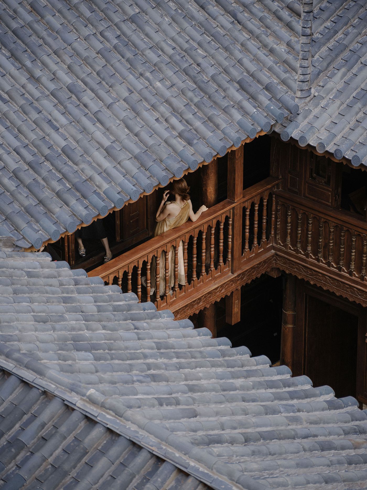 A woman in a flowing dress leans against a wooden railing, surrounded by traditional blue-tiled rooftops in an ancient courtyard. A woman in a flowing dress leans against a wooden railing, surrounded by traditional blue-tiled rooftops in an ancient courtyard.