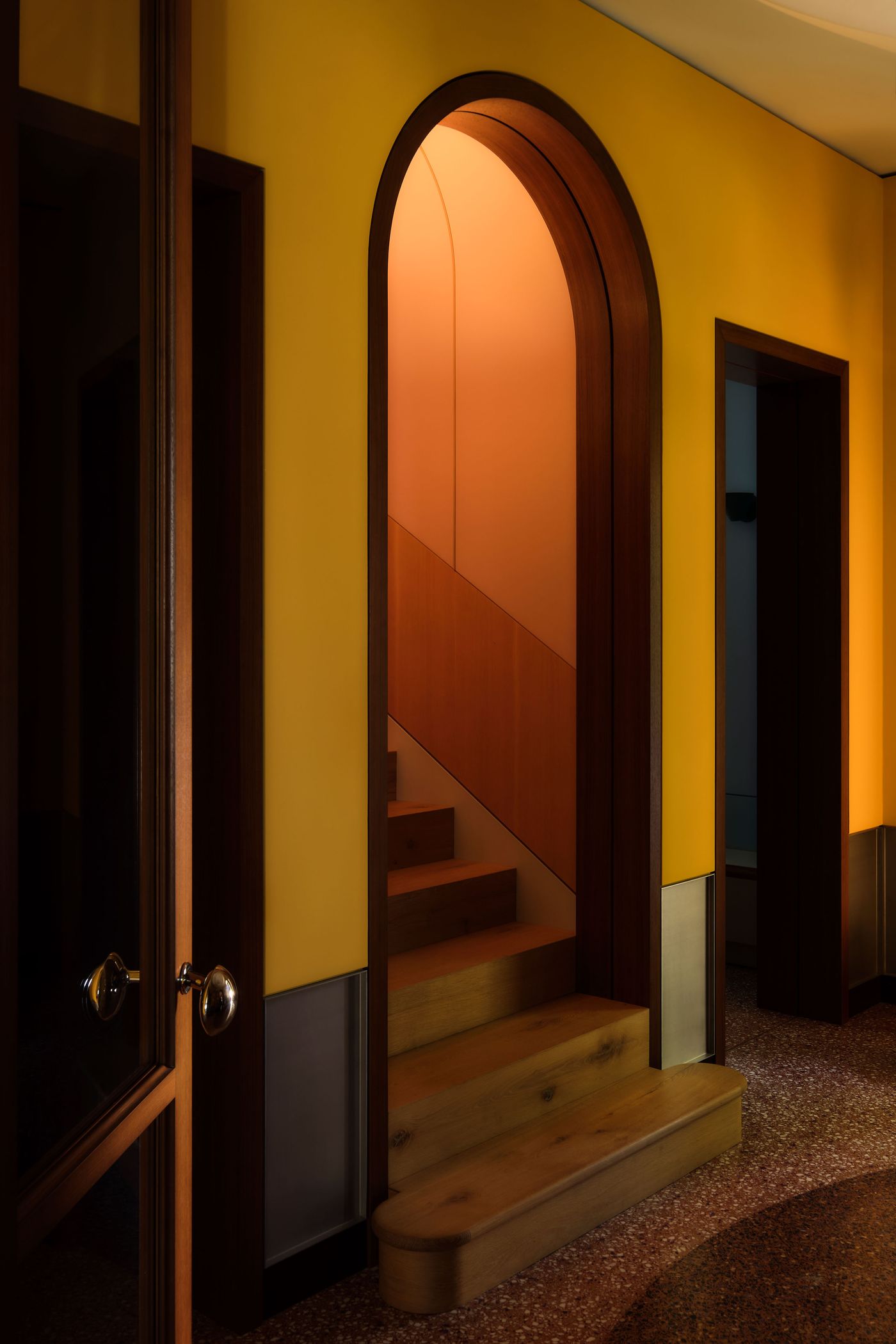A warmly lit hallway with a yellow wall and arched doorway leading to a wooden staircase, showcasing modern design elements.