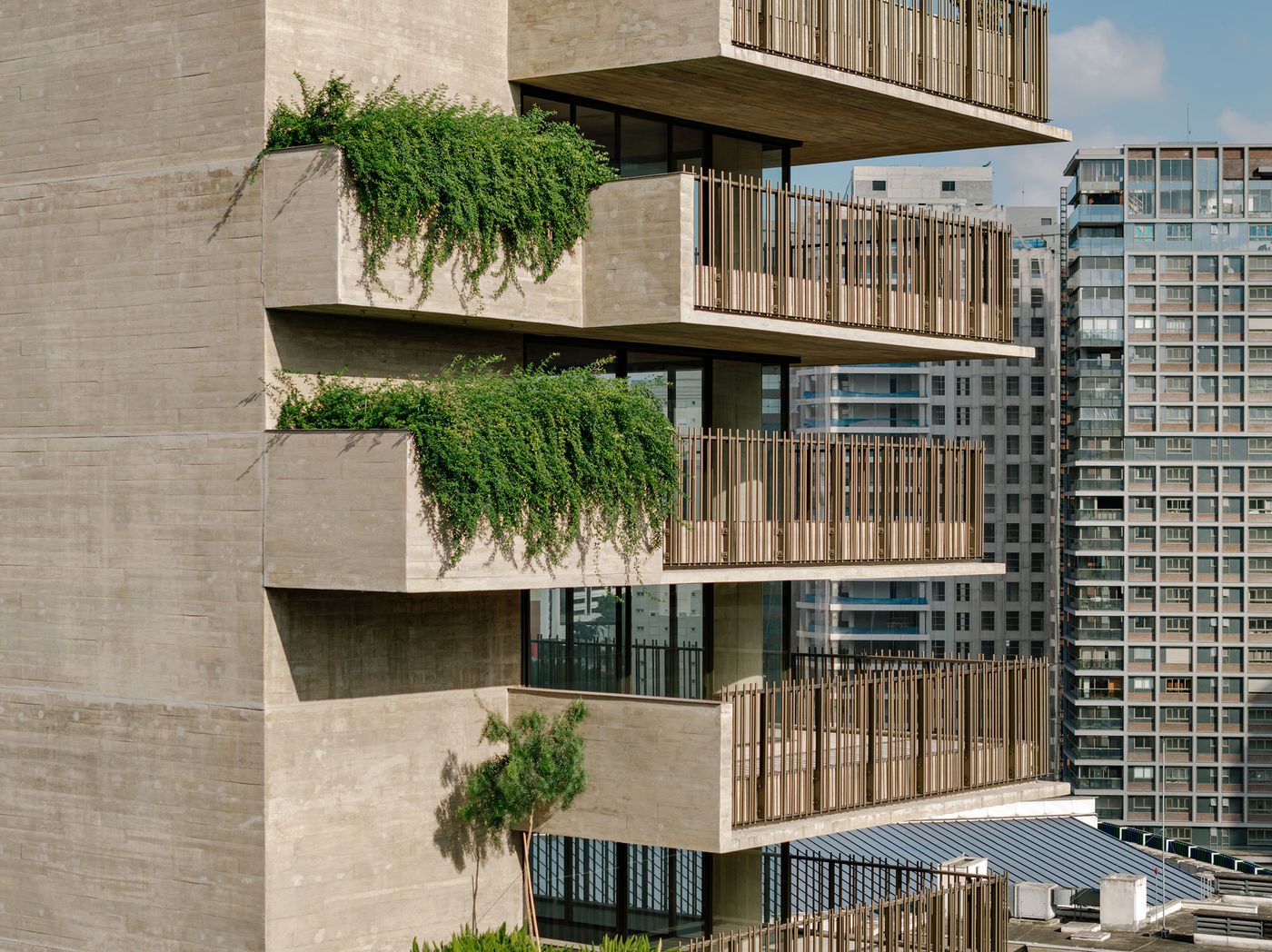 A close-up of the terraced concrete facade, highlighting sections where vibrant green plants spill over the edges, softening the rigid lines. The detailed vertical balustrades provide visual rhythm, emphasizing the building's commitment to biophilic design within its brutalist architectural style.