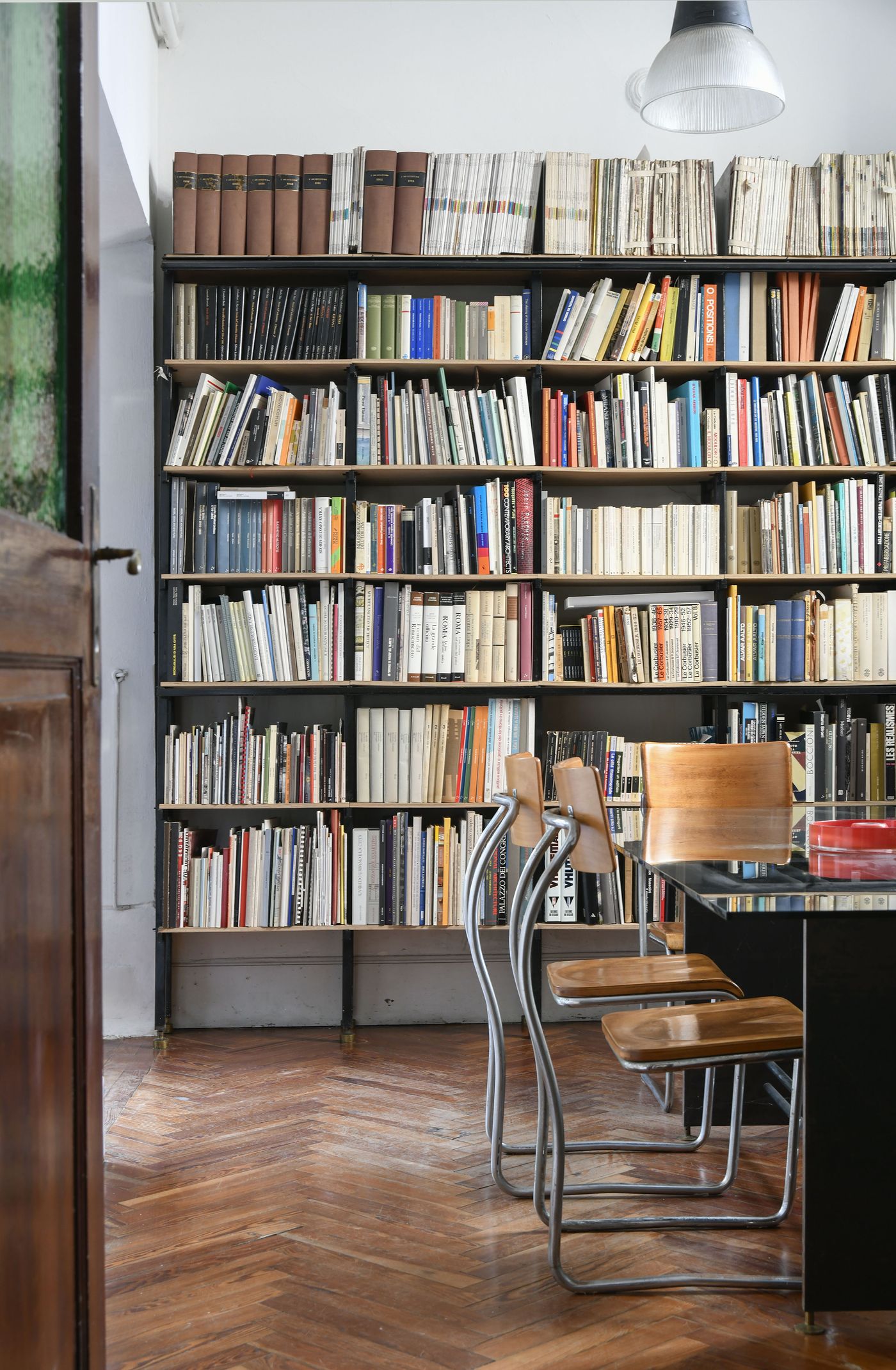 An extensive floor-to-ceiling bookshelf dominates the scene, filled with a diverse collection of books. In the foreground, a modern black table with a glass top is set with two sleek, curved metal chairs with wooden seats. The warm tones of the herringbone wooden floor complement the rich colors of the books and the polished surfaces of the furniture, creating a sophisticated and intellectual ambiance.