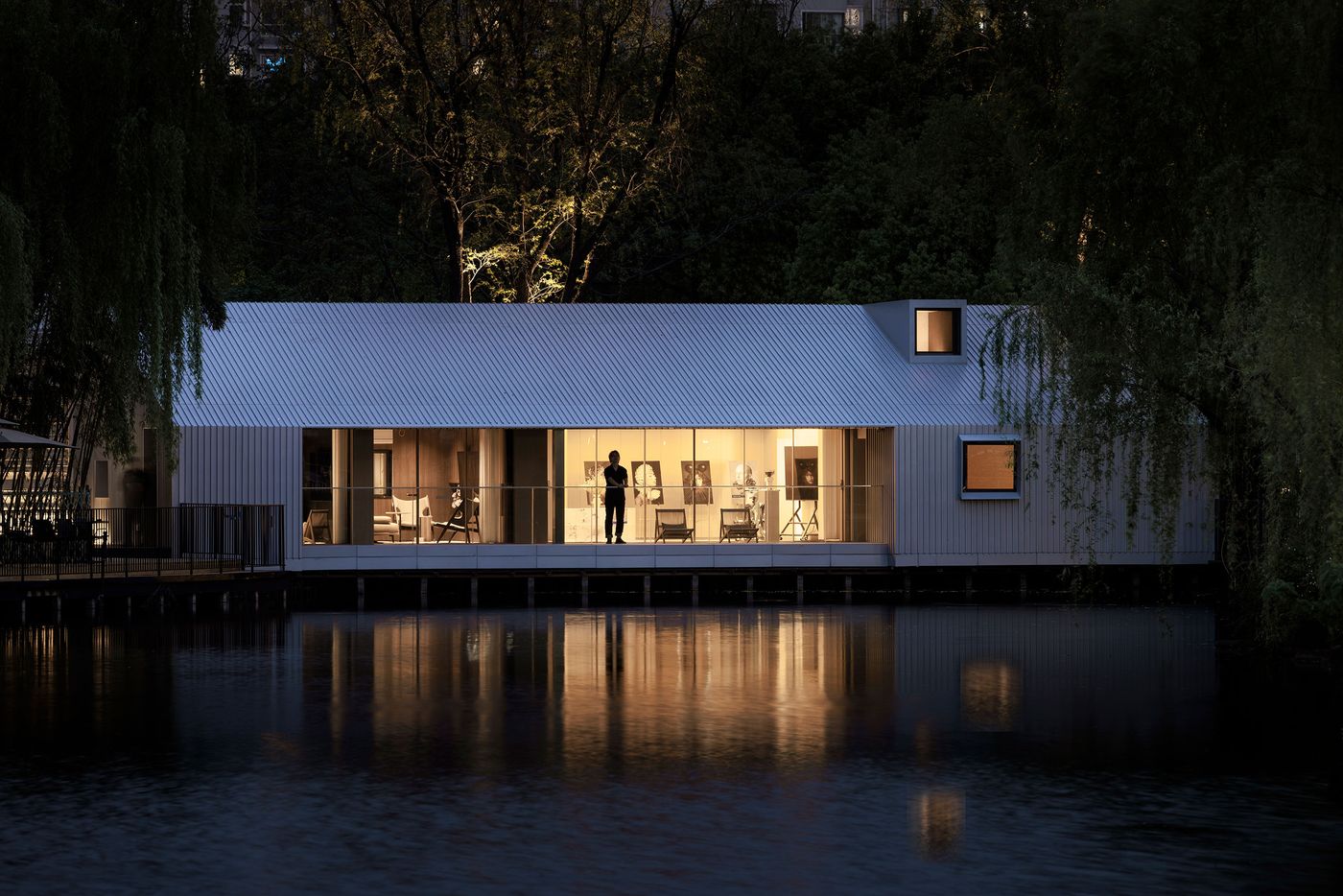 At twilight, Wutopia Lab's Lake House glows, its minimalist silver corrugated facade illuminated from within, casting a warm reflection on the still lake. A solitary figure stands silhouetted by the interior light, observing the serene scene. The design embodies modern tranquility, with dramatic lighting emphasizing the interplay between the interior’s warmth and the darkening external landscape. At twilight, Wutopia Lab's Lake House glows, its minimalist silver corrugated facade illuminated from within, casting a warm reflection on the still lake. A solitary figure stands silhouetted by the interior light, observing the serene scene. The design embodies modern tranquility, with dramatic lighting emphasizing the interplay between the interior’s warmth and the darkening external landscape.