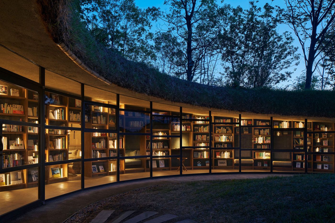 A night view of a modern library with glass walls, displaying illuminated bookshelves and a grassy roof, surrounded by trees. A night view of a modern library with glass walls, displaying illuminated bookshelves and a grassy roof, surrounded by trees.