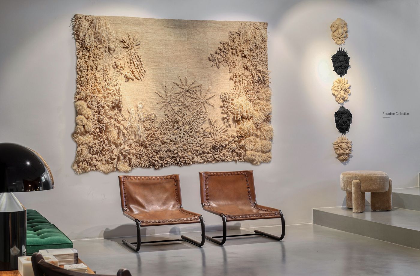 Inside the Estadeobruto showroom, a large, intricate textile art piece by Natalia Brilli Studio dominates the wall, featuring organic, textured forms in neutral tones. Below, two vintage-style lounge chairs crafted from dark leather and metal frames offer a relaxed counterpoint. A series of sculptural, textured mask-like pieces from the "Paradise Collection" are displayed vertically, adding an element of raw artistry. Inside the Estadeobruto showroom, a large, intricate textile art piece by Natalia Brilli Studio dominates the wall, featuring organic, textured forms in neutral tones. Below, two vintage-style lounge chairs crafted from dark leather and metal frames offer a relaxed counterpoint. A series of sculptural, textured mask-like pieces from the "Paradise Collection" are displayed vertically, adding an element of raw artistry.