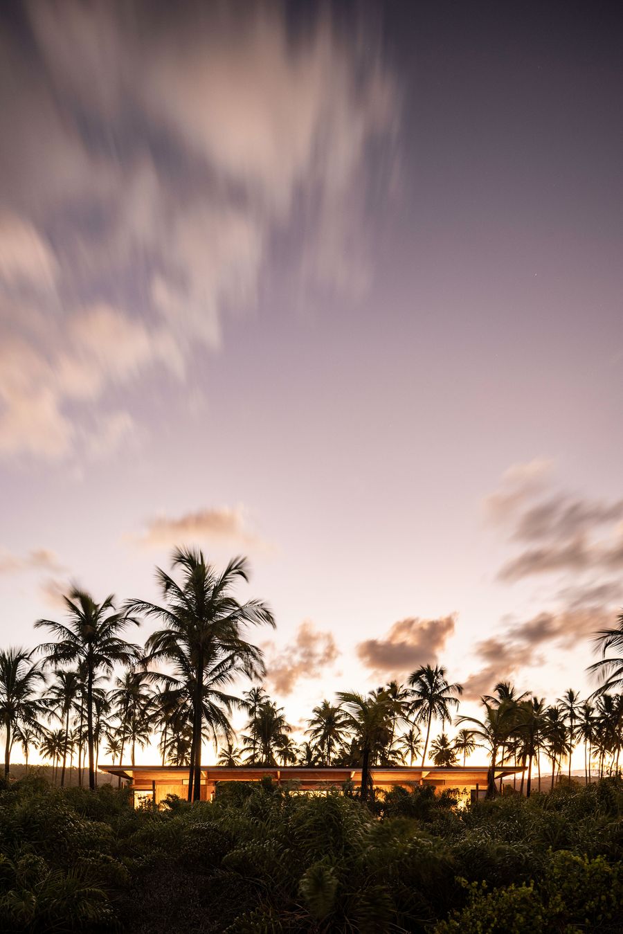 A low-slung, sunlit, low-slung structure nestles amidst dense tropical foliage and silhouetted palm trees under a dramatic, elongated sunset sky. The subtle glow from within hints at a serene, hidden retreat, emphasizing harmony with the natural environment. A low-slung, sunlit, low-slung structure nestles amidst dense tropical foliage and silhouetted palm trees under a dramatic, elongated sunset sky. The subtle glow from within hints at a serene, hidden retreat, emphasizing harmony with the natural environment.