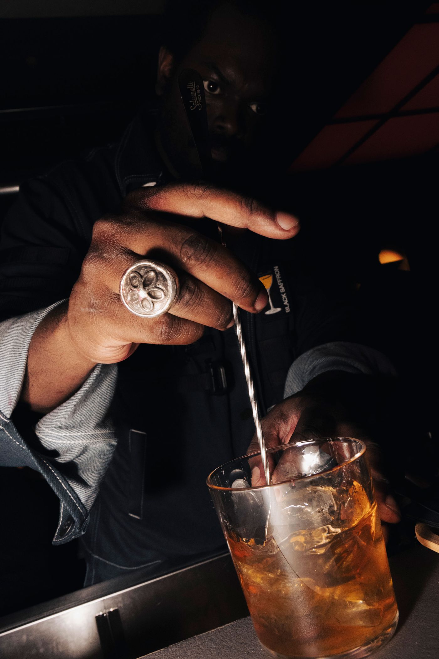 A bartender, wearing a silver ring, stirs a cocktail in a glass filled with ice in a dimly lit bar setting.