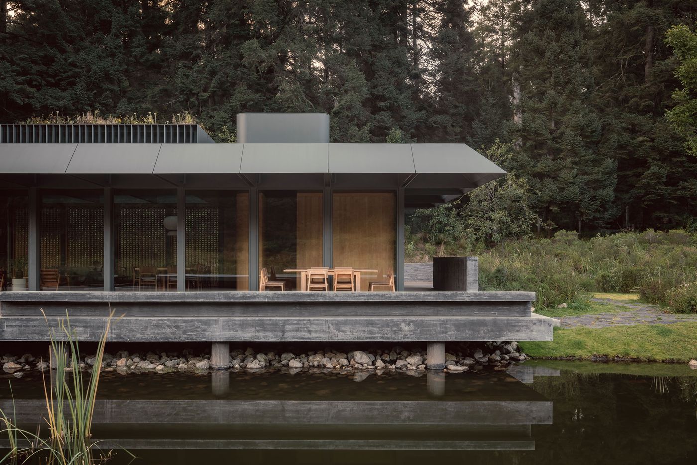 Modern house with large windows and a sloped roof, situated by a tranquil pond, surrounded by lush greenery and tall trees.