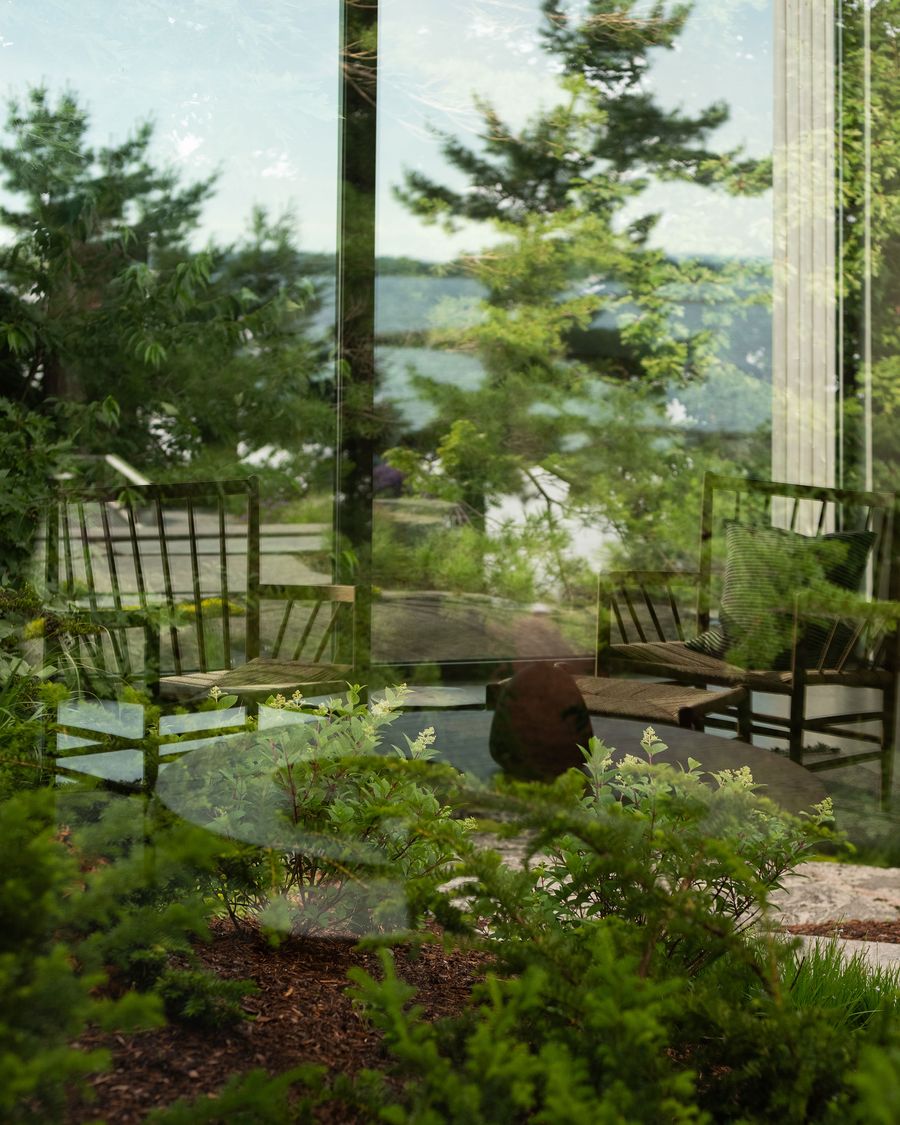 A serene view through a glass window, showcasing chairs, lush greenery, and a tranquil lake in the background. A serene view through a glass window, showcasing chairs, lush greenery, and a tranquil lake in the background.