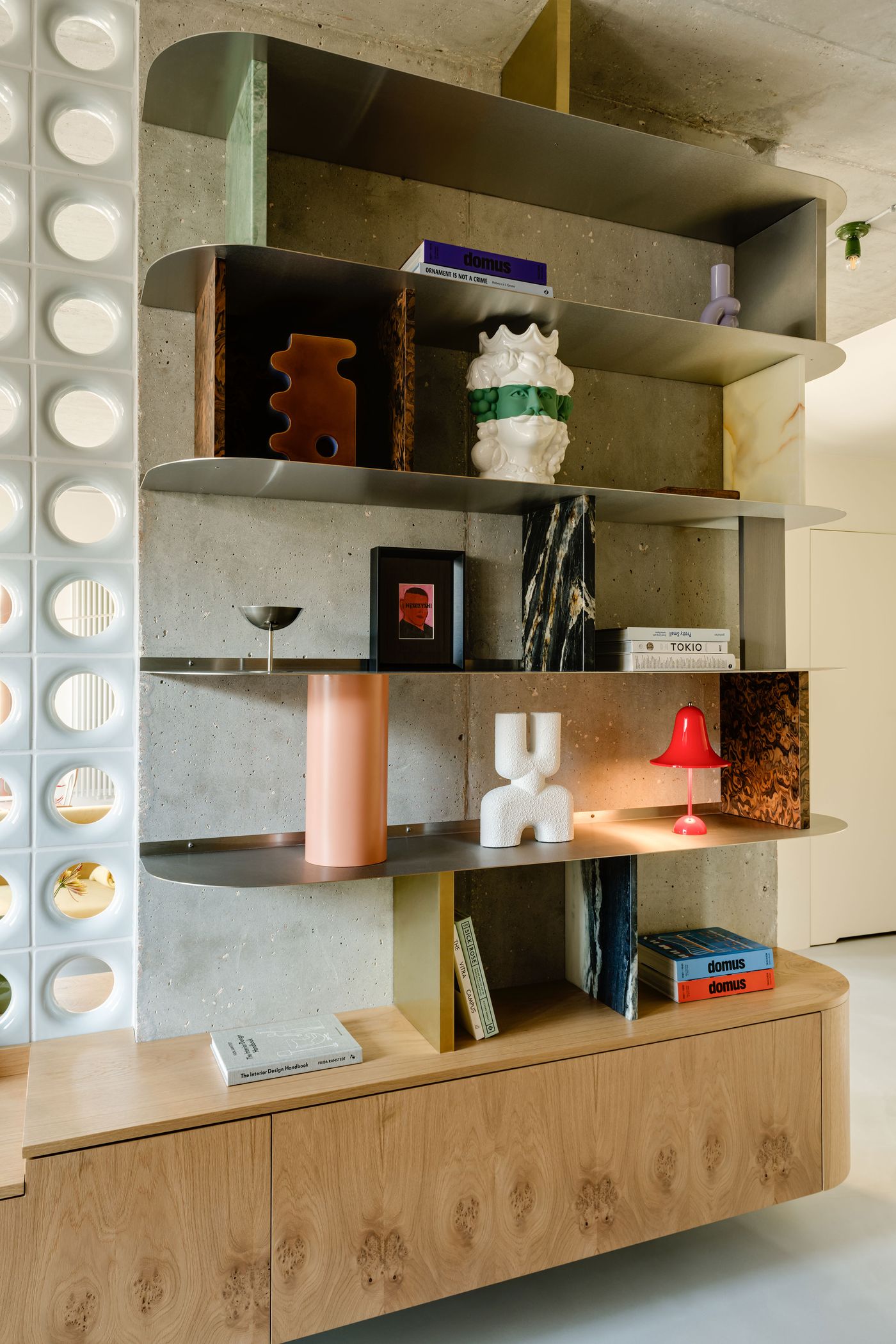 A custom open shelving unit mixes brutalist and mid-century cues with raw concrete, stainless steel, glossy wood, and marble dividers. Carefully curated objects—books, ceramics, and sculptural lighting—underscore a collector’s sensibility.