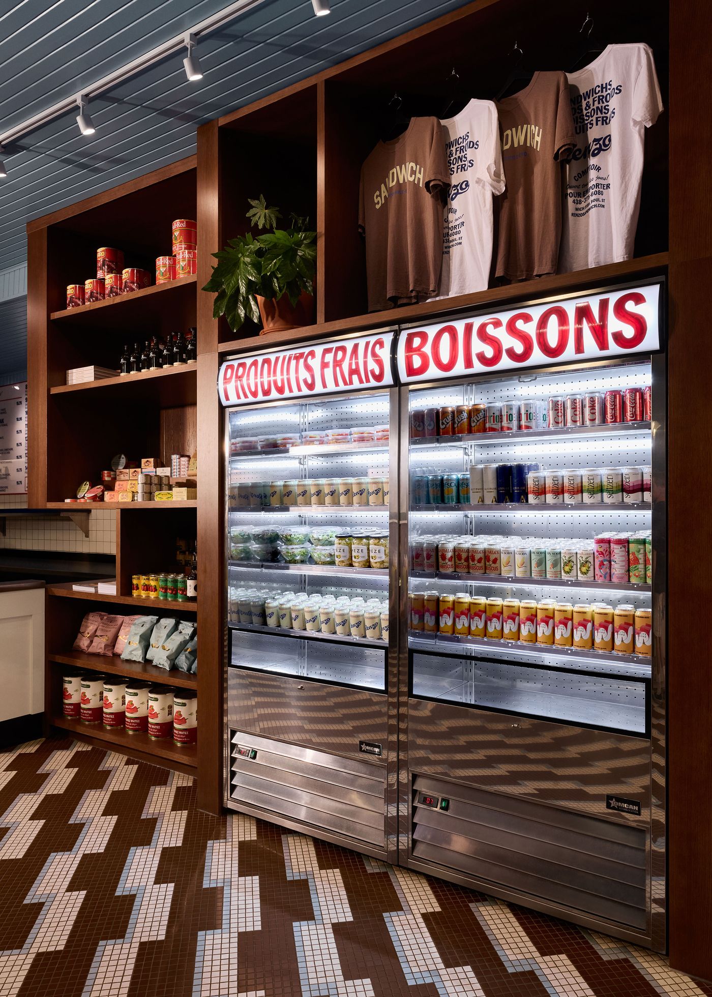 A well-stocked display of beverages and packaged goods fills a large shelving unit and two refrigerated units at Renzo Sandwich Shop. Above the coolers, signs in red lettering read "PRODUITS FRAIS" and "BOISSONS." Several t-shirts with the "Renzo Sandwich" logo are displayed on the top shelf, showcasing the brand's merchandise.