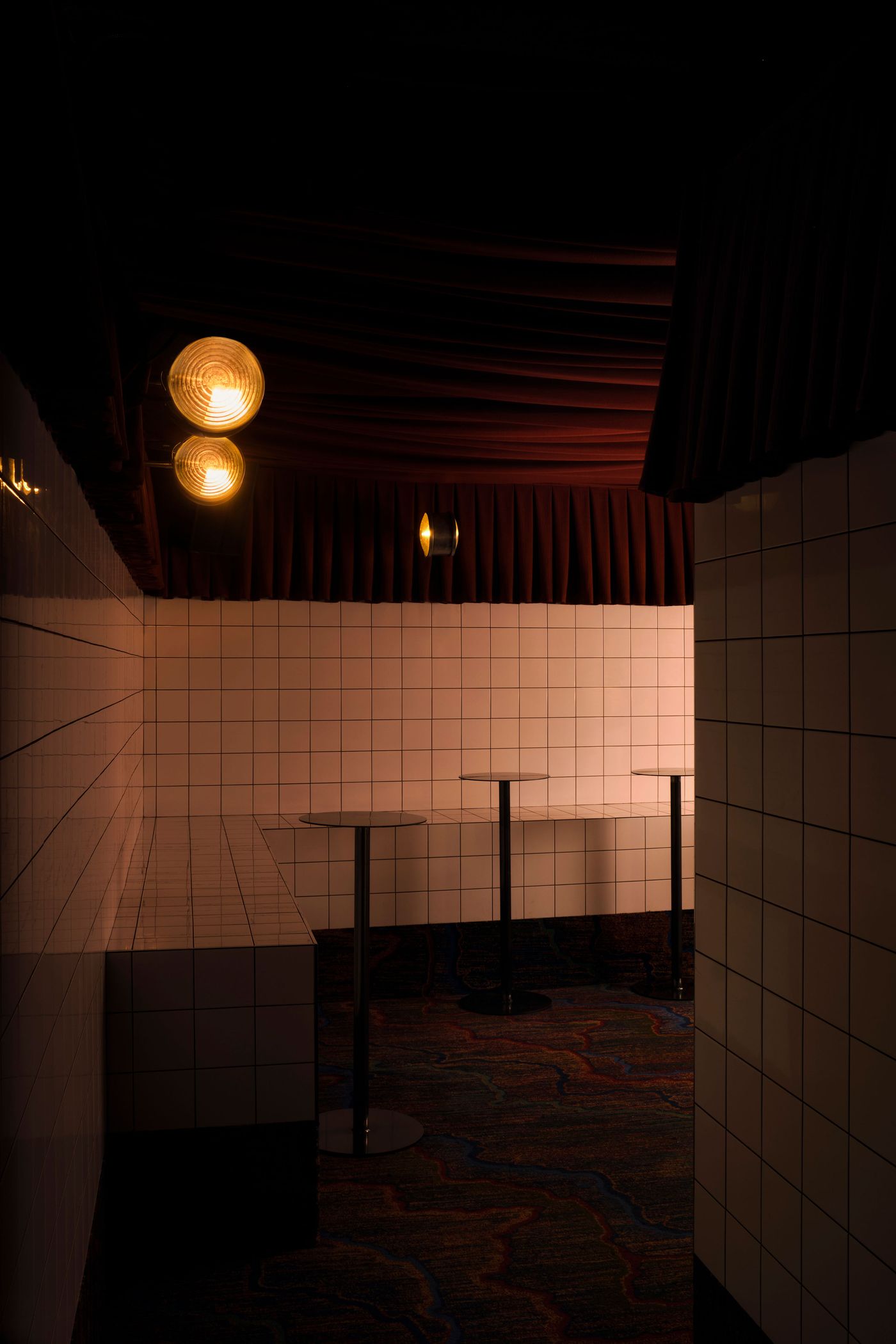 A striking composition within The Roses of Elagabalus, showcasing a patterned carpet leading towards a tiled wall. Two globular, orange-glowing ceiling lights illuminate a series of dark, tall bar tables, set against a backdrop of rich red pleated drapes. The interplay of textures and warm lighting creates a unique, artful, and intimate space.
